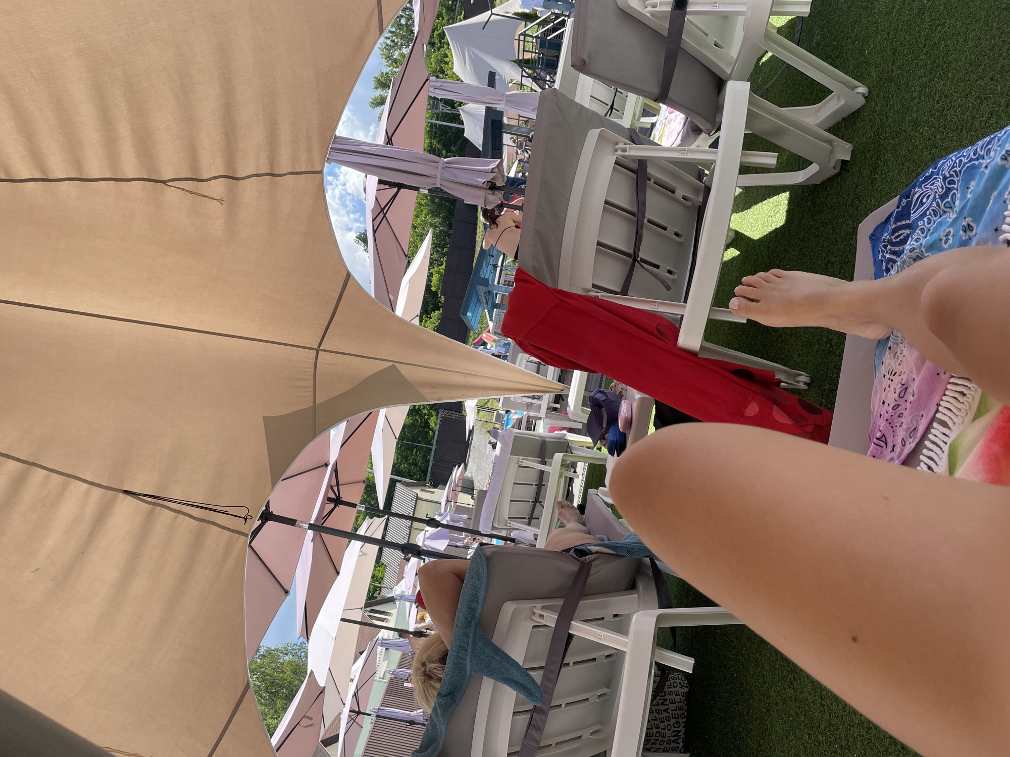 A first-person perspective shot shows a person's legs and feet resting on a lounge chair under a large tan canopy, at an outdoor pool area. The lounge chairs are white plastic with grey cushions, and the ground is covered in artificial grass. In the background, several pink and white umbrellas are set up, and people are seen relaxing and walking around. A blue towel is draped over one of the chairs. The environment suggests a sunny day at a resort or public pool. The location is identified as Zazymia, Ukraine.