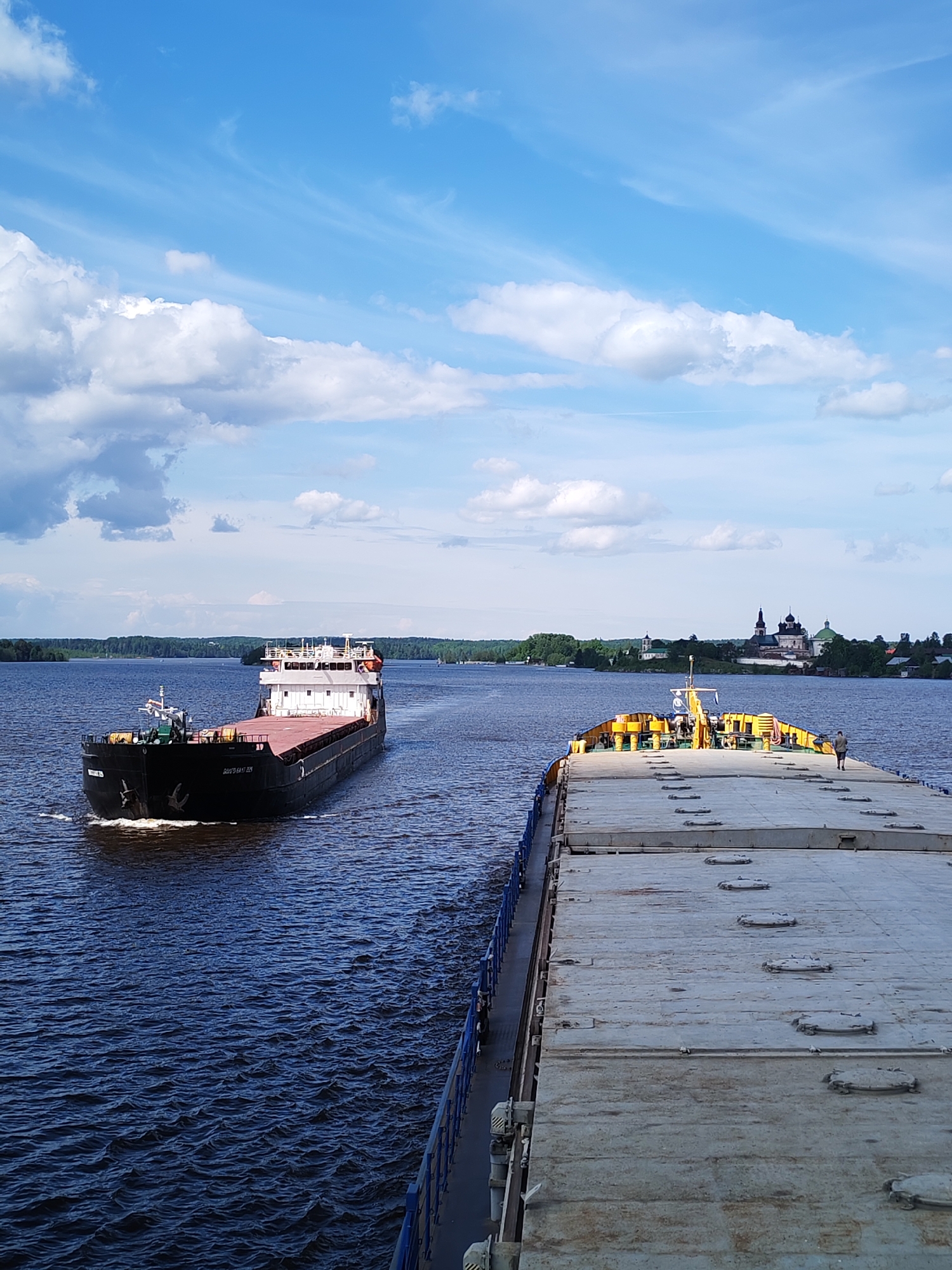 A daytime shot features two barges on a river, presumably the Volga River, near the town of Goritsy, Russia. One barge is traveling towards the viewer, while another moves away. A monastery stands on the shoreline. The sky is blue with scattered clouds.