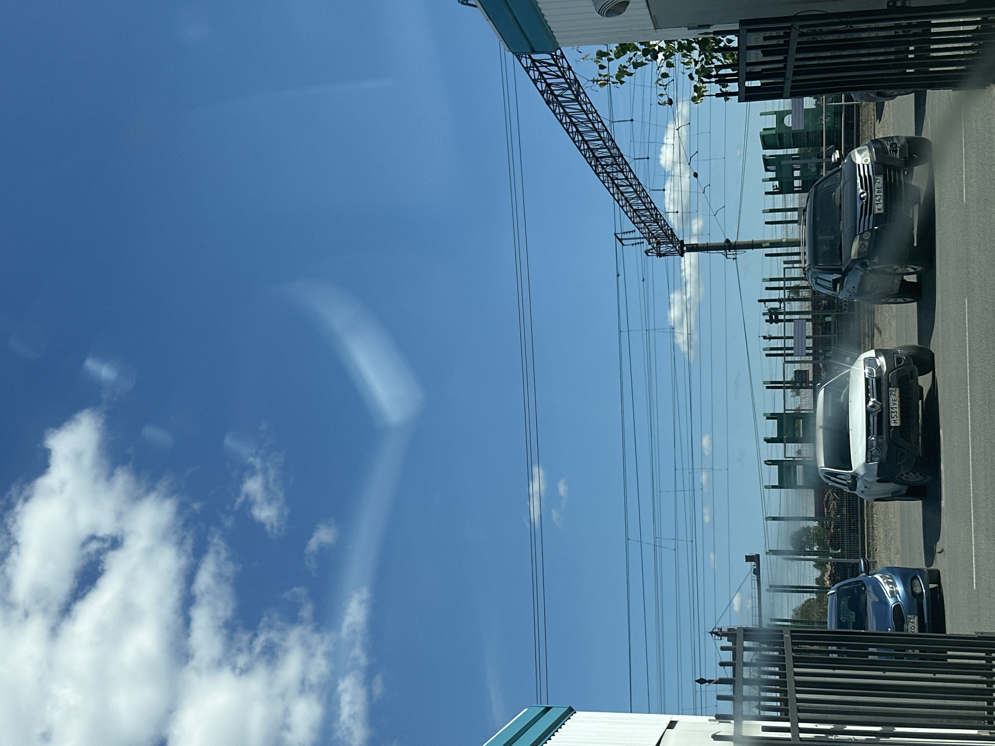 The image depicts an outdoor scene in Plesetsk, Russia, likely taken during daylight hours with a clear blue sky and scattered clouds. In the foreground, a black metal gate is visible, framing a view of a road and surrounding structures. Three vehicles are present on the road: a blue hatchback on the left, a white SUV in the center, and a black SUV on the right. The license plate of the white SUV reads "M531YE 29," and the license plate of the black SUV reads "K143BE 29." A small bird is standing on the asphalt between the blue and white vehicles.
In the midground, there are railway tracks and overhead power lines supported by poles. Beyond the tracks, a fence separates the area from a background of industrial-looking structures with green accents and ribbed metal walls. These structures appear to be part of an industrial facility or depot. The overall environment suggests a location near a railway line and an industrial zone within the city. The image appears to be taken from inside a vehicle, as evidenced by reflections on the surface in the upper portion of the image.