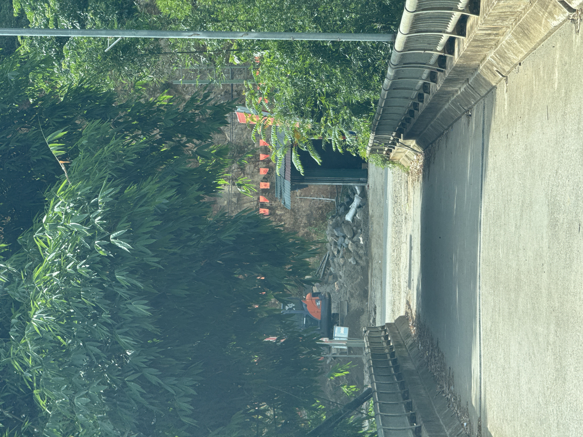 This image, captured from an elevated position likely a bridge or overpass, presents a vibrant, verdant scene under bright daylight, indicative of a sunny day. The perspective looks down onto a lower area, revealing both natural foliage and human construction.
In the foreground, a robust concrete bridge railing with a textured, possibly sound-dampening, design runs along the right side, separating the viewer from the scene below. Below the railing, a concrete wall or embankment descends, exhibiting signs of weathering. The left and central portions of the frame are dominated by dense, lush greenery, primarily consisting of bamboo and other leafy trees, with sunlight dappling through their leaves. A horizontal metal bar, possibly a utility pole component, spans the upper part of the image.
Further down and partially nestled among the trees, a building is visible, featuring a grey corrugated metal roof and a dark, open entrance. Adjacent to this structure, a construction or groundwork area is apparent, characterized by piles of rocks, debris, and some white sacks. An orange compact excavator is positioned in this area, suggesting ongoing earthwork or development.
A distinctive detail is a string of small, orange pennant flags, each bearing a yellow star, hanging near the building. These flags are a strong cultural indicator for Vietnam, aligning with the specified location context of Lao Chai. The scene captures a moment of active construction amidst a naturally rich environment. No discernible text is clearly visible on any signs or objects within the image.