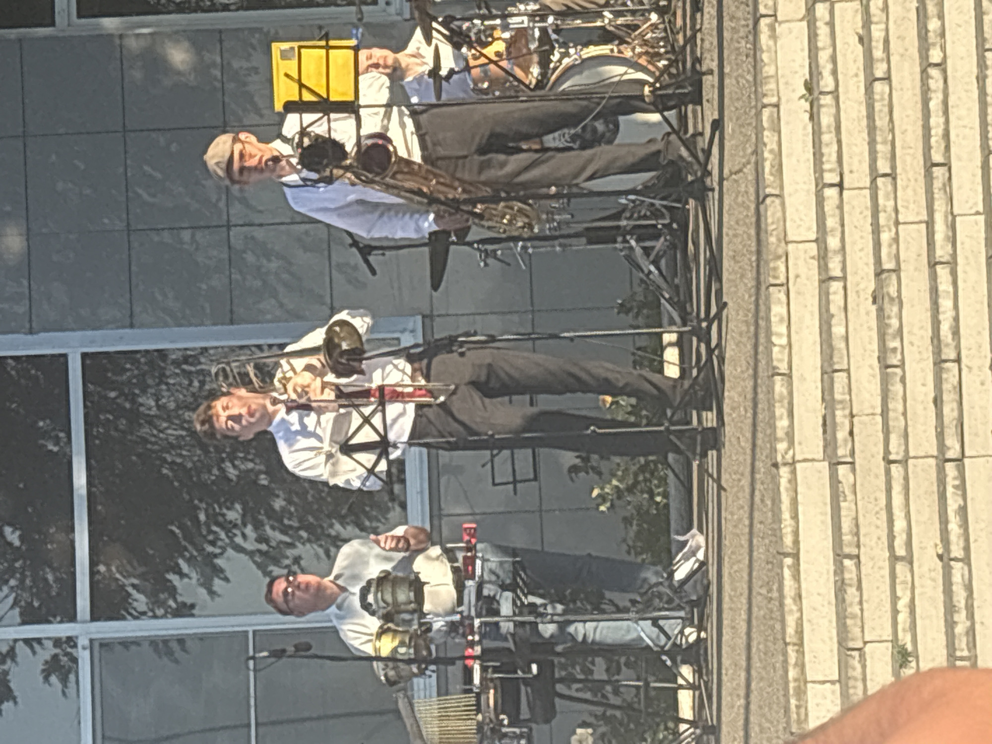 An outdoor performance is taking place on a raised stage, with a building facade of light gray tiles and large windows in the background. Four musicians are visible. On the left, a man wearing a light blue shirt and jeans is playing percussion instruments, including bongos and a marimba-like instrument. He is wearing sunglasses. In the center, a man in a white shirt and dark pants plays a trombone. To his right, another man in a white shirt and dark pants, wearing a flat cap and glasses, plays a saxophone. Behind the saxophonist and slightly to the right, a fourth musician is seated behind a drum kit. The musicians appear to be engaged in a lively performance. The environment suggests a sunny day.