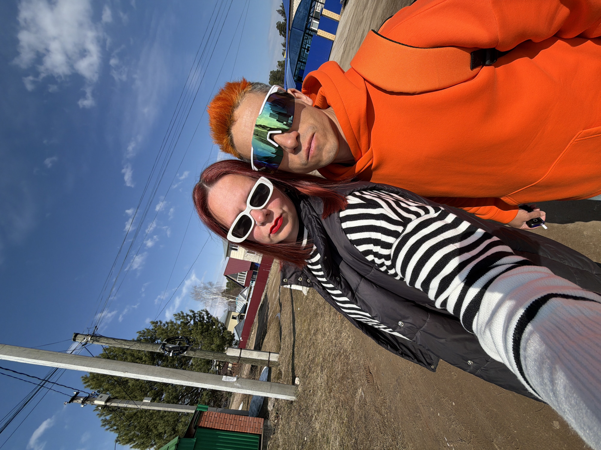A bright, sunny selfie captures two individuals outdoors, likely in Chaikovsky, Russia. The image is taken from a slightly upward angle, showing a vast expanse of clear blue sky with sparse white clouds.
The person in the foreground on the right is a male with vibrant orange, spiky hair and large, white-framed mirrored sunglasses that reflect the sky and surrounding environment. He wears a bright orange hoodie and looks directly at the camera with a neutral expression. His right hand is partially visible, holding a small object, possibly an electronic cigarette or vape device.
The person on the left is a female with red hair, wearing white-framed rectangular sunglasses and bold red lipstick. She is dressed in a black and white striped turtleneck sweater layered under a black puffer vest. Her expression is also neutral, looking directly at the camera.
The background suggests a rural or semi-urban environment. To the left, several utility poles with power lines stretch across the frame, leading towards the horizon. Beyond the poles, there are residential buildings with distinct sloped roofs and fences, some appearing red and green. The ground is dry, covered in brown grass or dirt, indicating late winter or early spring. In the distance, evergreen trees are visible, and further back, a large blue and white industrial-style building is partially seen. The bright sunlight casts soft shadows, confirming it's daytime. No visible text is clearly discernible in the image.