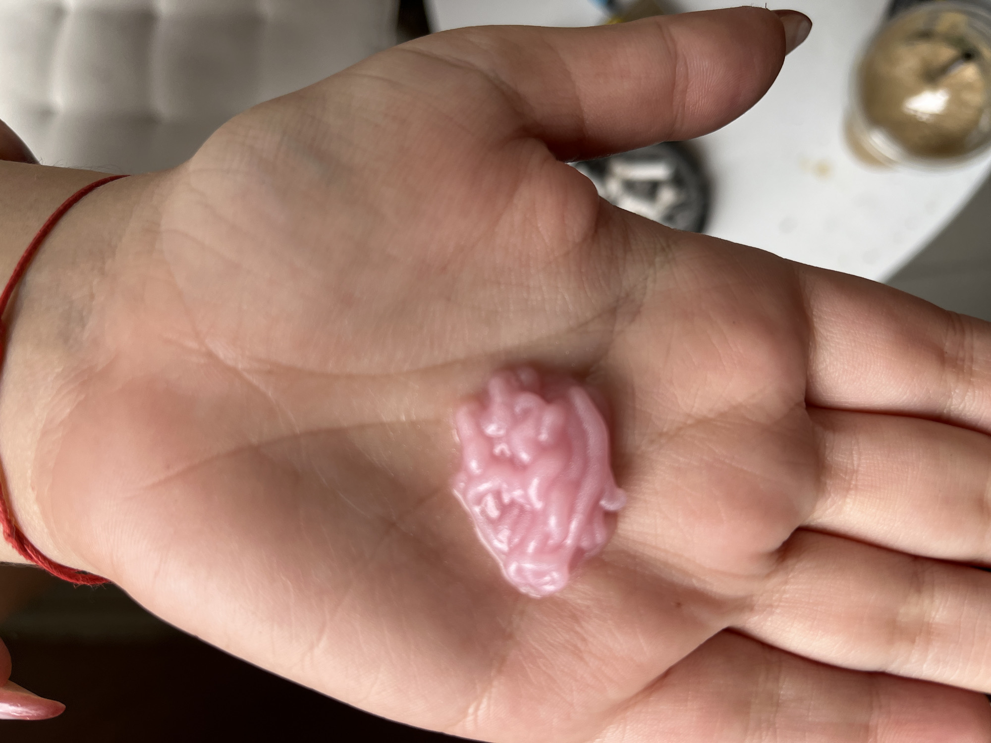 A close-up, overhead shot shows a person's left palm holding a small, pink, brain-shaped object. The object appears to be made of a soft, pliable material, possibly candy or a stress toy, with intricate folds and textures resembling a human brain. The palm is fair-skinned with visible lines and creases. A thin red string is visible wrapped around the person's wrist. The background is blurred, with hints of a white, tufted surface and a clear glass containing a light brown liquid, suggesting an indoor setting. There are no discernible landmarks indicating the city of Sofia, Bulgaria.