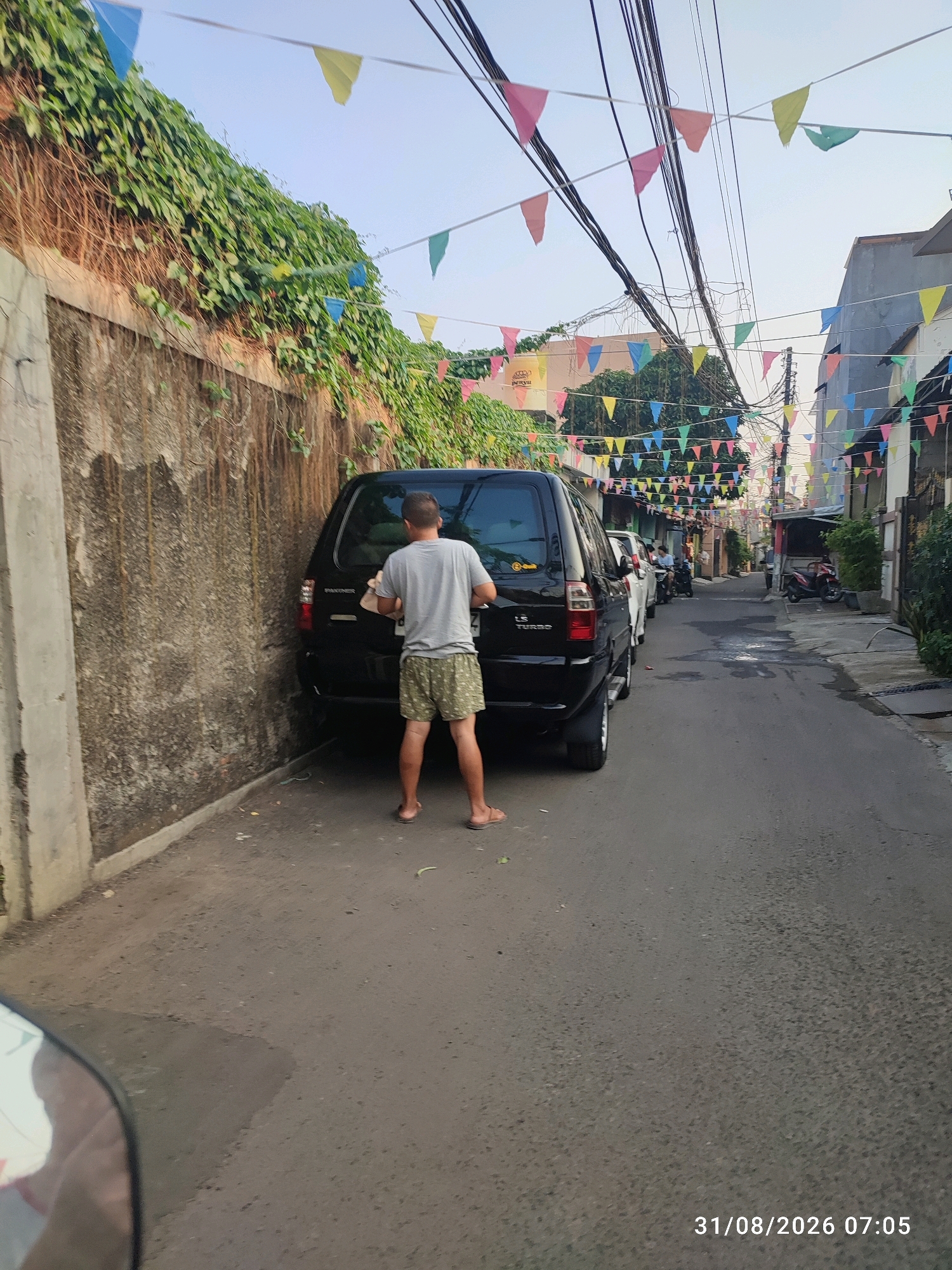 The image captures a narrow street in East Jakarta, Indonesia, adorned with colorful triangular flags strung across power lines. A man in a gray t-shirt and patterned shorts stands with his back to the camera, facing a black SUV parked on the left side of the street. He appears to be cleaning or interacting with the rear of the vehicle, holding a light-colored object in his right hand. The street is paved with asphalt and shows signs of recent rain or moisture. To the left of the man and the SUV is a weathered, textured wall covered in dense green ivy. Other vehicles, including cars and motorcycles, are visible further down the street, hinting at a residential or commercial area. The overall scene suggests a typical day in a bustling Indonesian neighborhood, possibly during a local festival or celebration given the festive decorations. The timestamp "31/08/2026 07:05" is visible in the lower right corner.