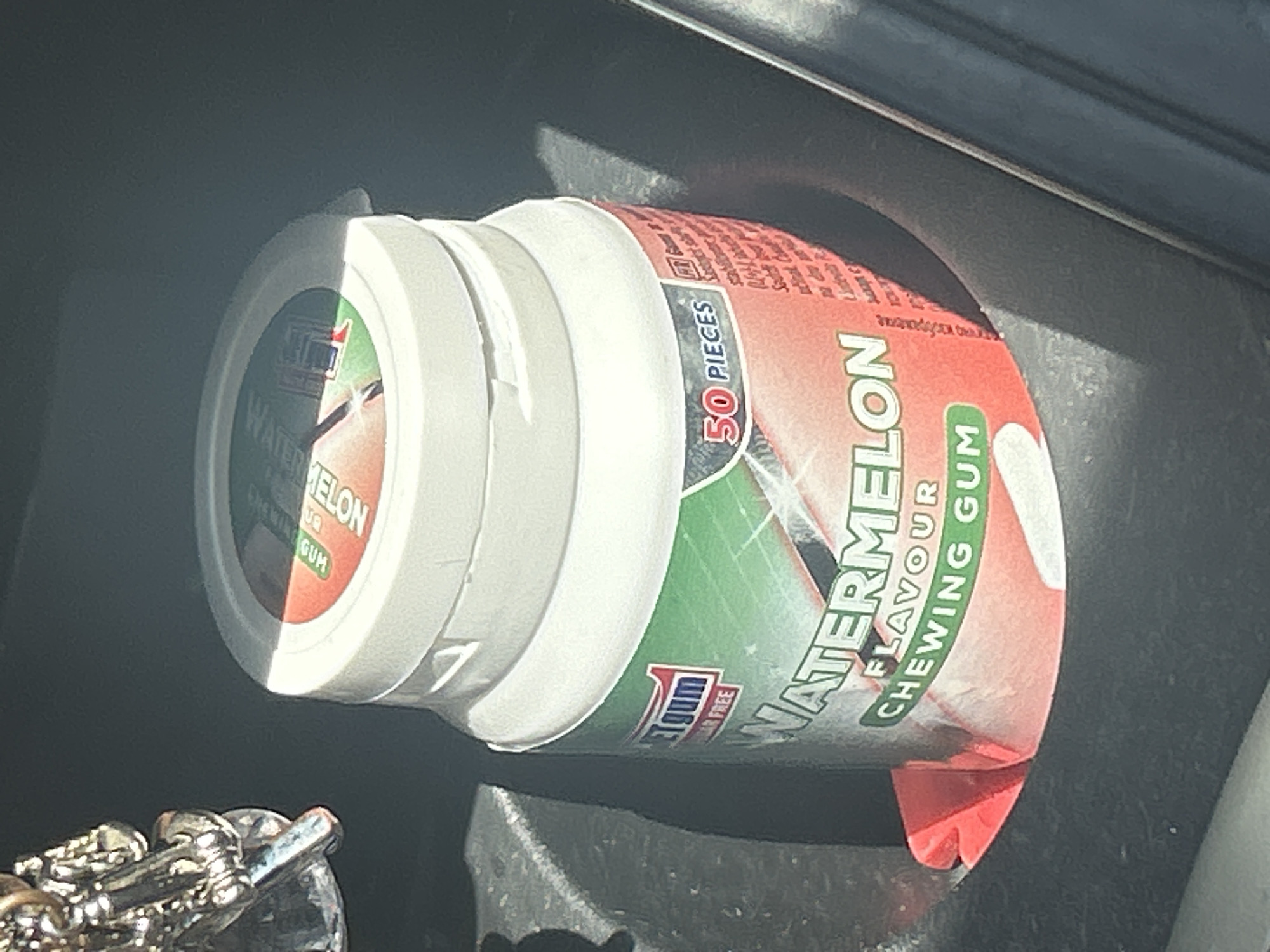 The image shows a close-up of a white plastic container of "WATERMELON FLAVOUR CHEWING GUM". The container is round with a white lid. The label is predominantly red and green, featuring a stylized depiction of a watermelon slice. Visible text includes "WATERMELON FLAVOUR", "CHEWING GUM", "50 PIECES", and "SUGAR FREE". The container appears to be placed in a dark, recessed area, possibly a car's cup holder or storage compartment. The lighting suggests it is daytime, with sunlight casting shadows and highlights on the container. A portion of a silver-colored metal object, possibly jewelry, is visible in the lower left corner of the image. The setting is likely Plovdiv, Bulgaria.