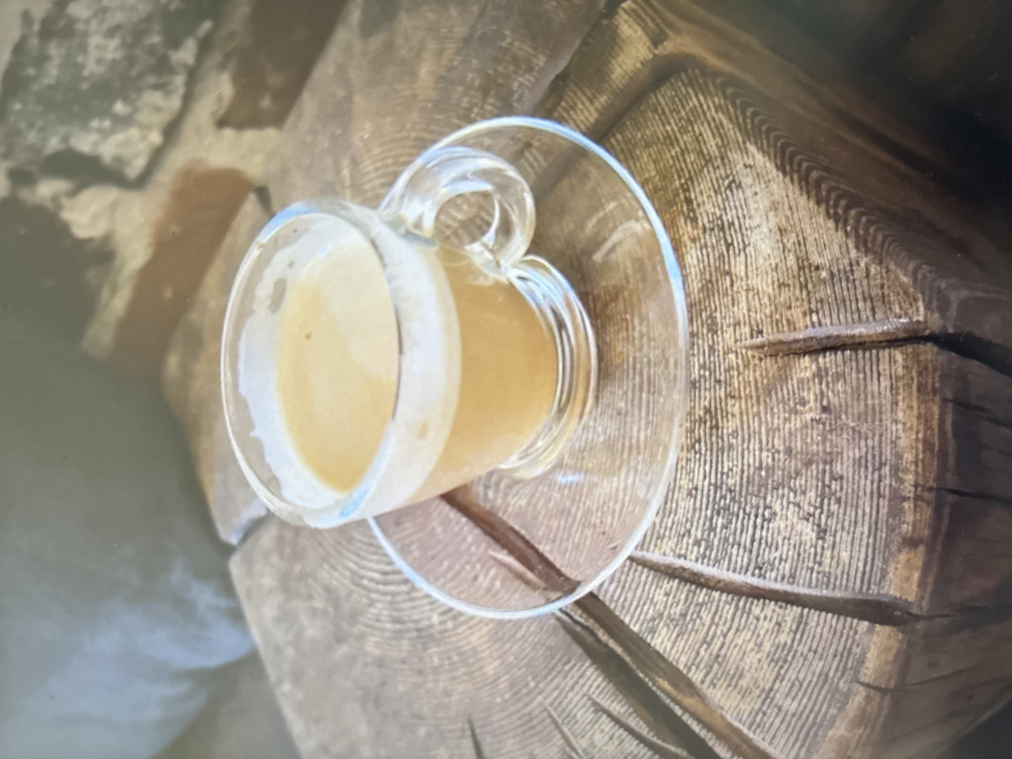A clear glass cup filled with a light brown liquid, likely coffee or tea, sits on a matching clear glass saucer. The saucer rests on a rustic wooden surface with prominent grain patterns and visible cracks. In the background, out of focus, are what appear to be rough stone or brickwork and a soft, blurred object, possibly upholstery. The lighting suggests an indoor setting. This image is not located in Novosibirsk, Russia.
