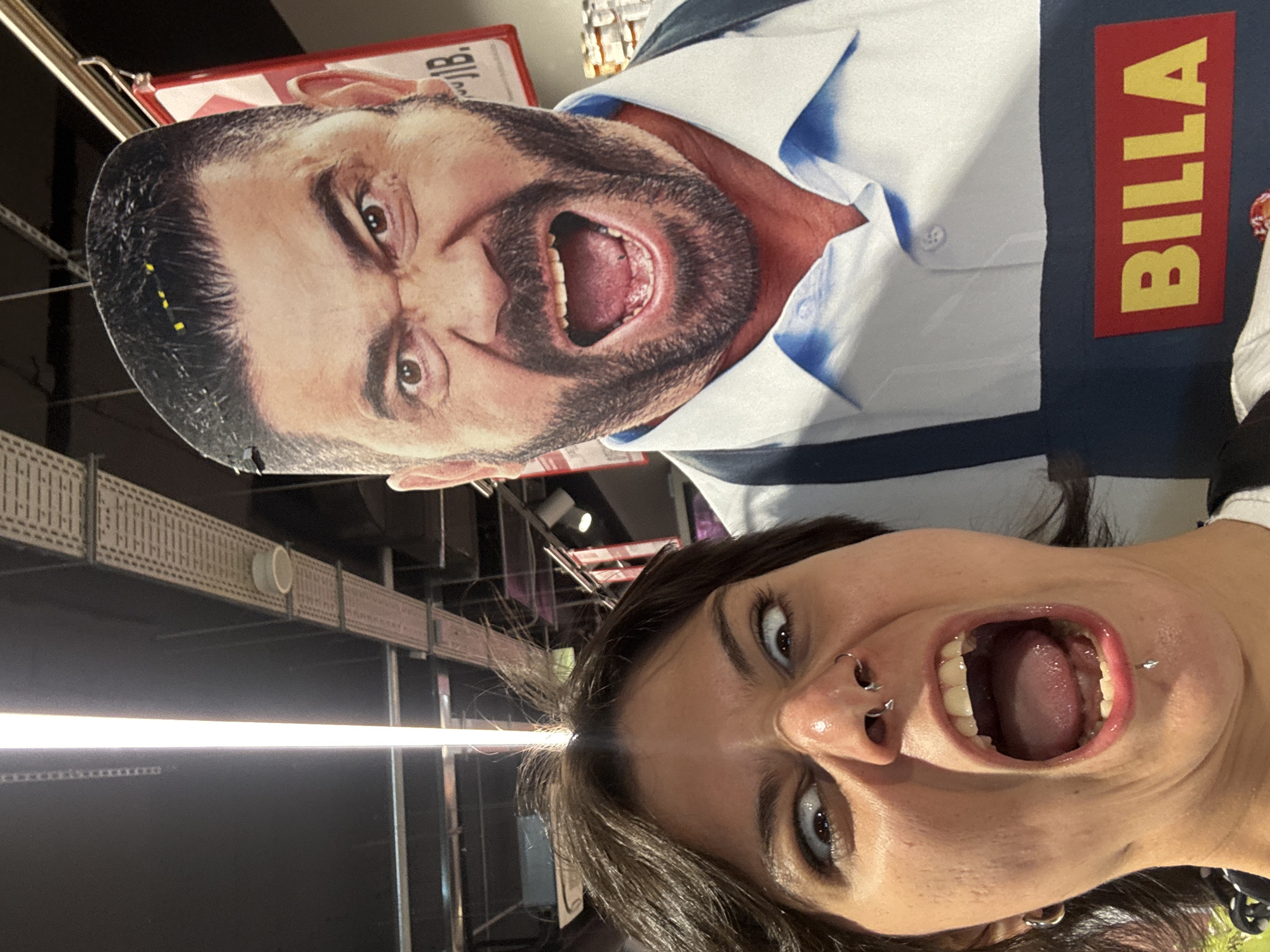 The image depicts two people, one is a woman with brown hair, facial piercings and dark eye makeup, smiling with her mouth open. The second is a flat cutout of a man with a dark beard and open mouth, wearing a white and blue shirt with the word "BILLA" in yellow letters on a red rectangle. The background shows an indoor environment that could be a supermarket. I do not have the information to identify the location as Sofia, Bulgaria.