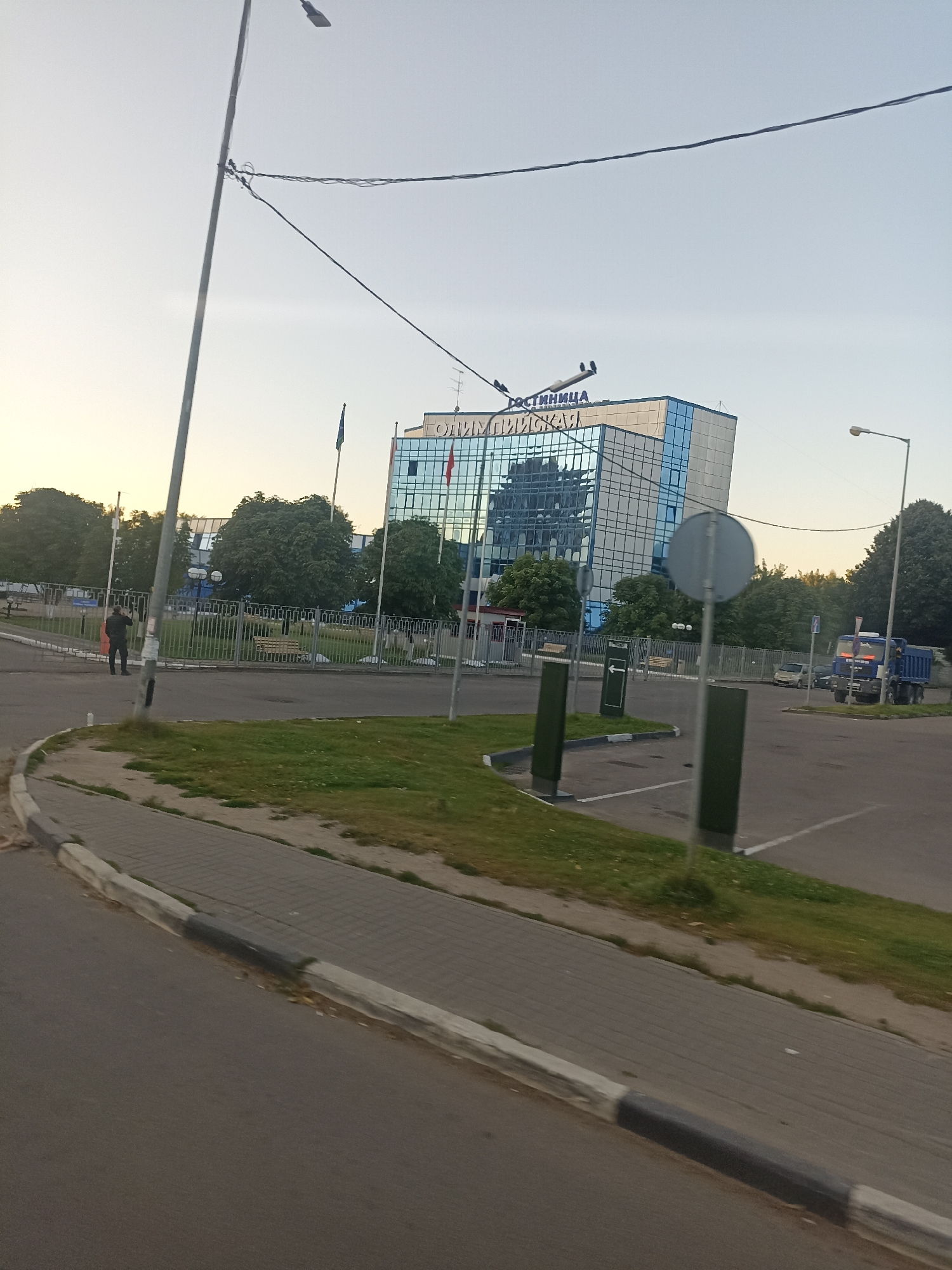 The image shows a street scene in Chekhov, Russia, with a modern glass-fronted building in the background. The building has "ГОСТИННИЦА" (Gostinnitsa, meaning Hotel) written in large blue letters across its top. A person in dark clothing is walking away from the camera towards a metal fence. A blue truck is parked to the right of the frame. The foreground features a paved sidewalk and road, with a grassy area to the left. Trees and streetlights are visible throughout the scene. The sky is overcast.