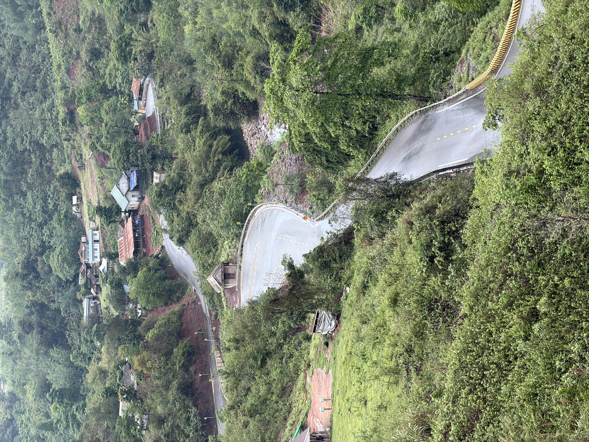 The image shows a section of road winding through a mountainous, densely vegetated area in La Lay Commune, Vietnam. The road is wet, suggesting recent rain, and features yellow lane markings and guardrails. Several dwellings are visible along the lower section of the road, indicating a residential area. The terrain is steep, with vegetation covering most of the hillsides. A partially covered structure is visible near the edge of the hill.