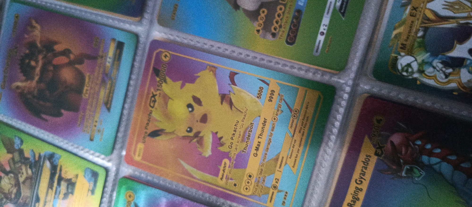 The image shows a close-up view of a Pokémon card binder containing several holographic Pokémon cards. The cards are arranged in plastic sleeves. In the center of the frame is a "Go Pikachu" GX card, which features a yellow Pikachu with a lightning bolt. To its left is a card with a dragon-like Pokémon. To the right of the "Go Pikachu" card is a card featuring "M Rayquaza EX". The lighting suggests an indoor environment, possibly a room with artificial light. The binder and cards appear to be in good condition. The image does not contain any discernible information related to the city of Brwinów, Poland.
