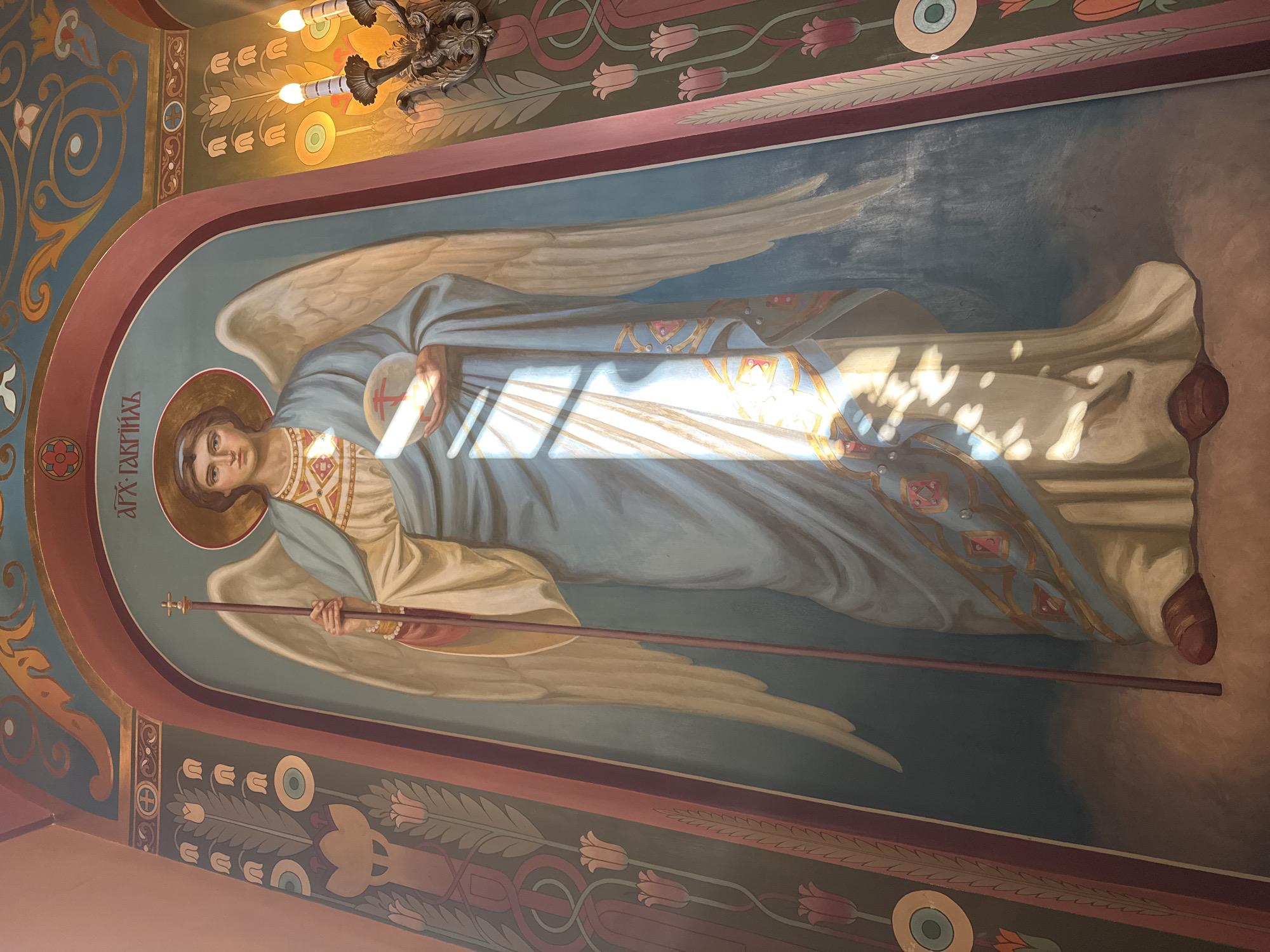 The image is a detail of an Orthodox mural likely inside a church or cathedral. It depicts an angel figure painted in traditional style, with wings, a halo, and holding a staff. The style suggests it may be located in Saint Petersburg, Russia, given the prevalence of Orthodox churches with similar murals in the city. The specific location and identity of the angel cannot be determined without more information. The painting is illuminated by sunlight.