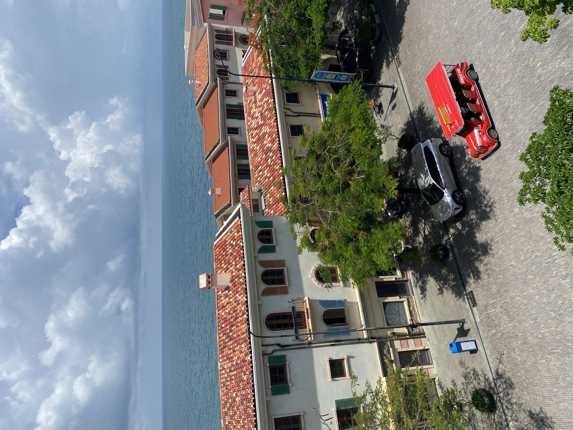 The image depicts a scene in Phú Quốc, Vietnam, featuring Mediterranean-style architecture with terracotta tiled roofs overlooking a calm blue sea under a cloudy sky. In the foreground, a cobblestone street is visible, with a red open-top shuttle bus labeled "Buggy Service" and a silver SUV parked on it. Lush green trees cast shadows on the street. The buildings have light-colored facades, arched windows, and green shutters. The overall impression is a resort-like environment with a coastal view.