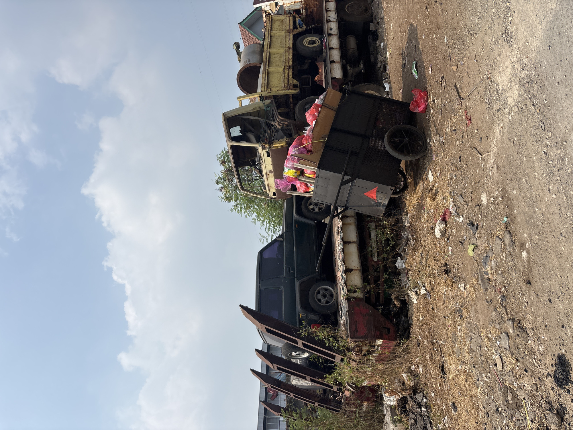 The image shows a junkyard in Bekasi, Indonesia, with two trucks and a dark SUV parked on a flatbed trailer. The first truck, a yellow flatbed, is loaded with a large concrete pipe. The second truck, also yellow, appears to be older and is partially dismantled. A black cart filled with colorful bags is positioned in front of the trucks. The ground is covered in dry grass, dirt, and scattered debris, suggesting an outdoor, unmaintained area. The sky is blue with scattered white clouds.