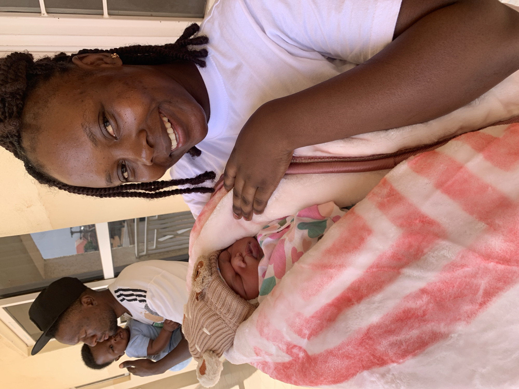 A medium shot depicts a smiling woman holding a swaddled baby outdoors. The woman, who is Black, wears a white t-shirt and has braided hair with a gold earring visible. The baby is wrapped in a pink and white striped blanket and wears a tan knit hat. In the background, a man in a black baseball cap holds another baby. The location is identified as Jos, Nigeria. The background features a building facade with a window and a beige painted wall.