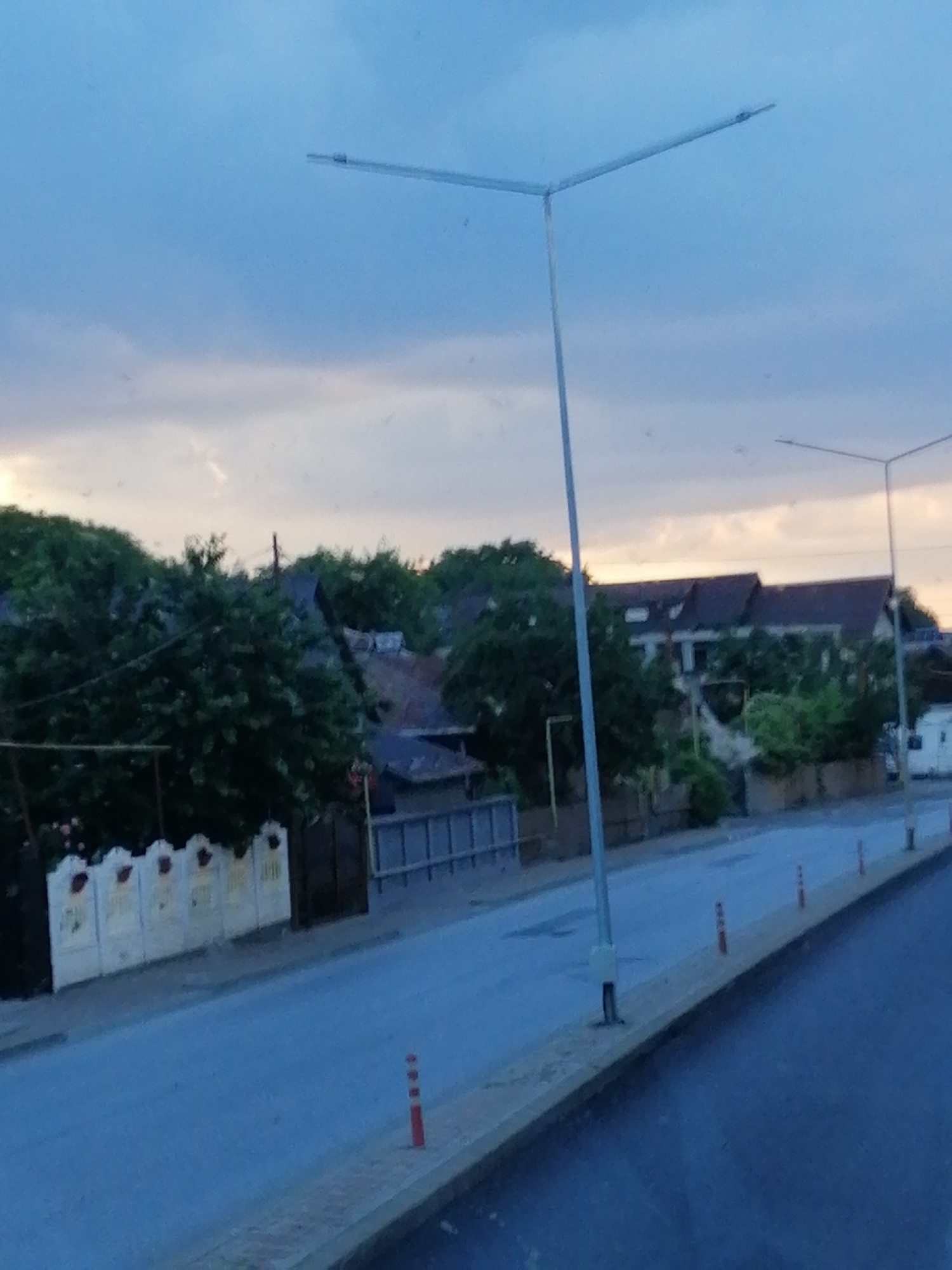 A wide shot captures a street scene at dusk, possibly in a suburban or rural area, given the presence of houses and dense greenery. The sky is overcast with hues of blue and soft orange, suggesting the sun has recently set. The foreground features a road with a light blue surface, bordered by a yellowish curb. Several orange and white traffic cones are strategically placed along the curb, marking a boundary.
On the left side of the road, a white fence with decorative elements is visible, partially obscured by lush green trees and bushes. Behind the fence, the roofs of several houses peek through the foliage, with some architectural details like chimneys and dormer windows discernible. A tall, modern street lamp with a Y-shaped arm stands prominently in the middle ground, its metal structure stark against the sky. Another similar street lamp is visible in the distance on the right.
Further down the street, more houses with dark roofs and some residential buildings with visible windows can be seen, interspersed with trees. A white recreational vehicle is parked on the right side of the road in the background. The overall impression is one of a quiet street at the end of the day, with elements of both residential life and essential infrastructure.
