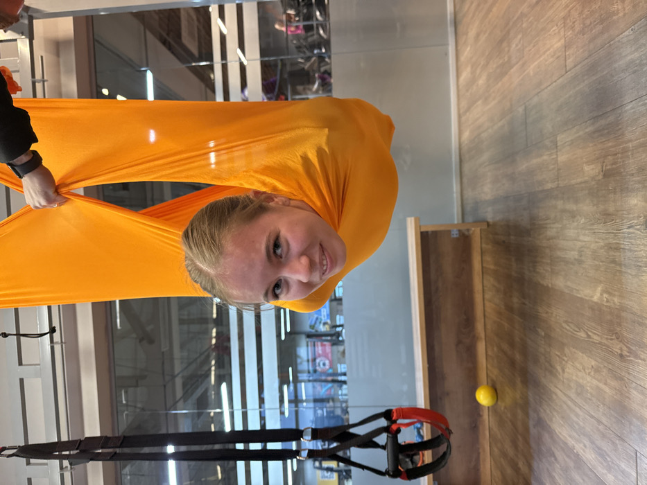 An indoor scene in a fitness facility located in Chaikovsky, Russia, depicts a young female suspended within a bright orange aerial yoga hammock. Her head, featuring light brown hair and a smiling expression, protrudes from the fabric, directed towards the viewer. A partial view of an individual's right hand, wearing a black wristband, holds the orange fabric from above, suggesting support or adjustment. To the left of the frame, a black TRX suspension trainer with red foot cradles hangs vertically. In the mid-ground, a light brown wooden bench extends along a wall. The background consists of a large glass partition revealing a wider gym area with blurry figures and exercise equipment. A small yellow spherical object rests on the light wood-effect laminate flooring. The overall environment is well-lit and appears modern.