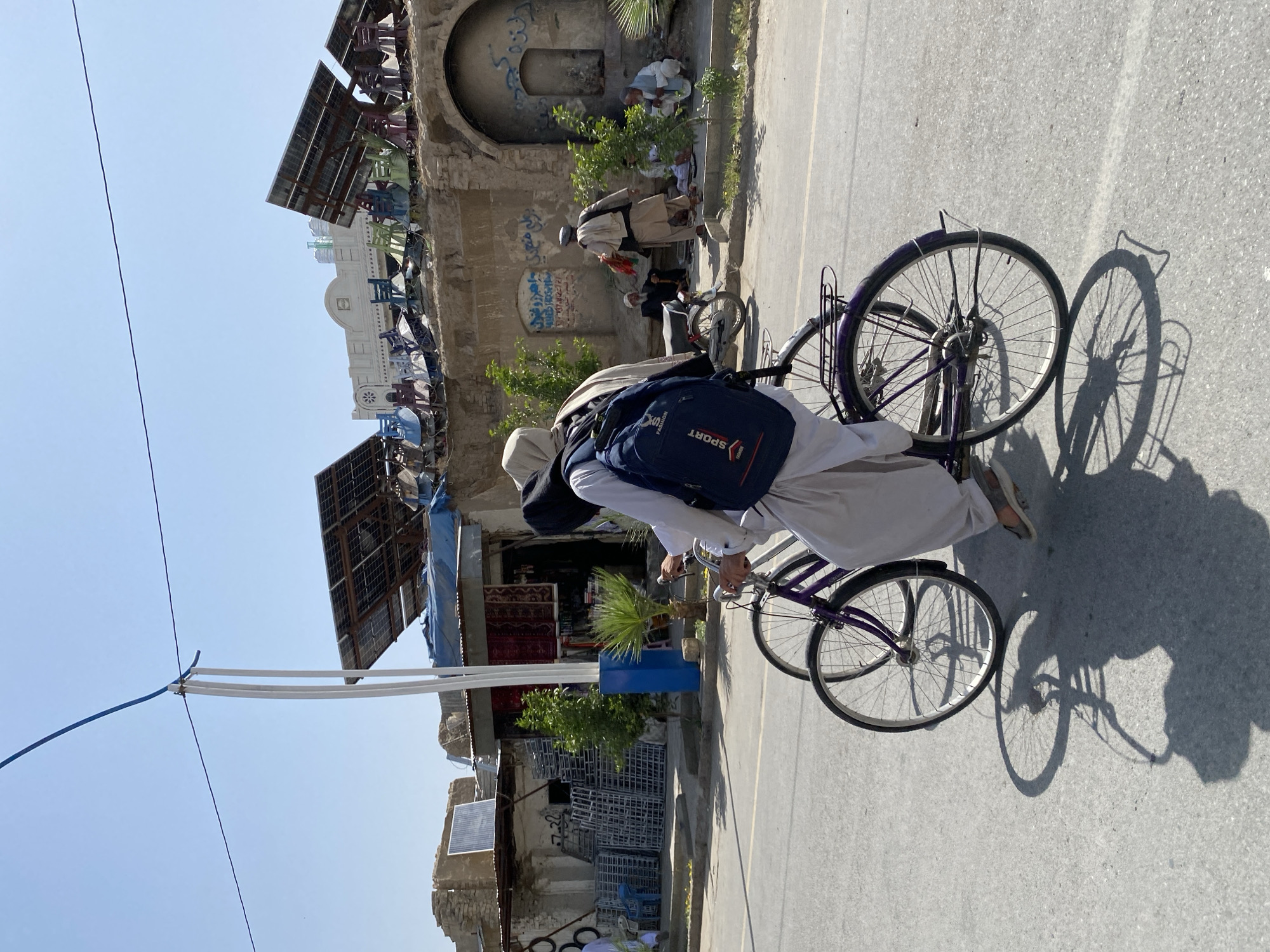 In Kandahar, Afghanistan, two men are riding a tandem bicycle on a paved street under a clear blue sky. The man in front is wearing a light-colored headscarf and traditional shalwar kameez, with a dark blue backpack. The man behind him is wearing a similar outfit. The bicycle is purple. In the background, buildings are visible, some with exposed brick and others with tin roofs. Solar panels are mounted on several structures. Some people are sitting on a ledge, and a motorcycle is parked nearby. Various items are displayed outside a shop, including what appear to be carpets and metal racks. A blue and white pole stands near the street. The overall scene depicts a street view in a city with a mix of old and new infrastructure, and signs of daily life.