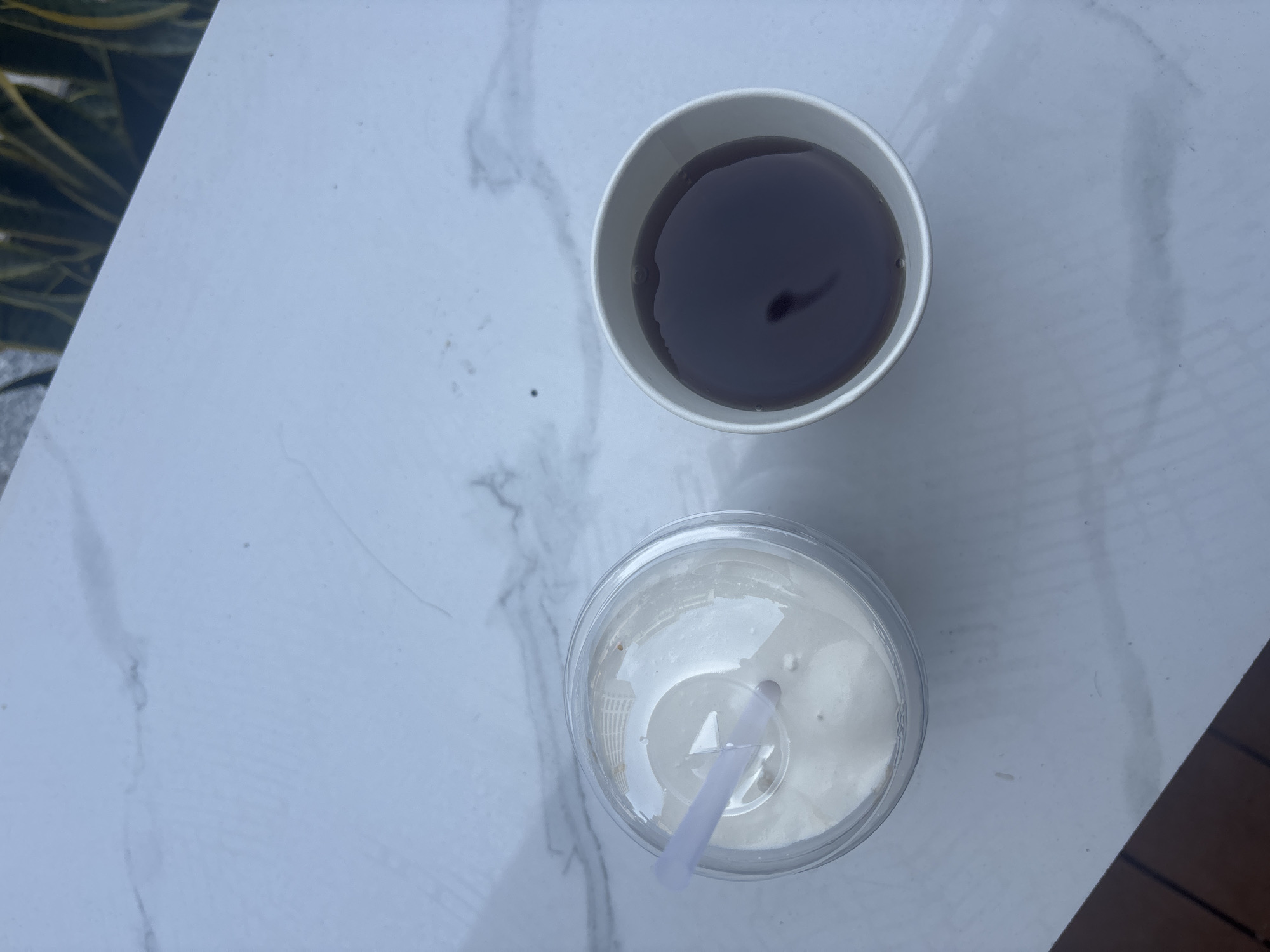 Two beverages are placed on a white marble-patterned surface. On the left is a clear plastic cup filled with a light-colored, opaque liquid, likely a smoothie or milkshake, with a white straw. The cup has a clear plastic lid with a hole for the straw. On the right is a white paper cup filled with a dark brown liquid, presumably coffee or tea. The background shows a blurry out-of-focus view of green foliage and a gray textured surface.