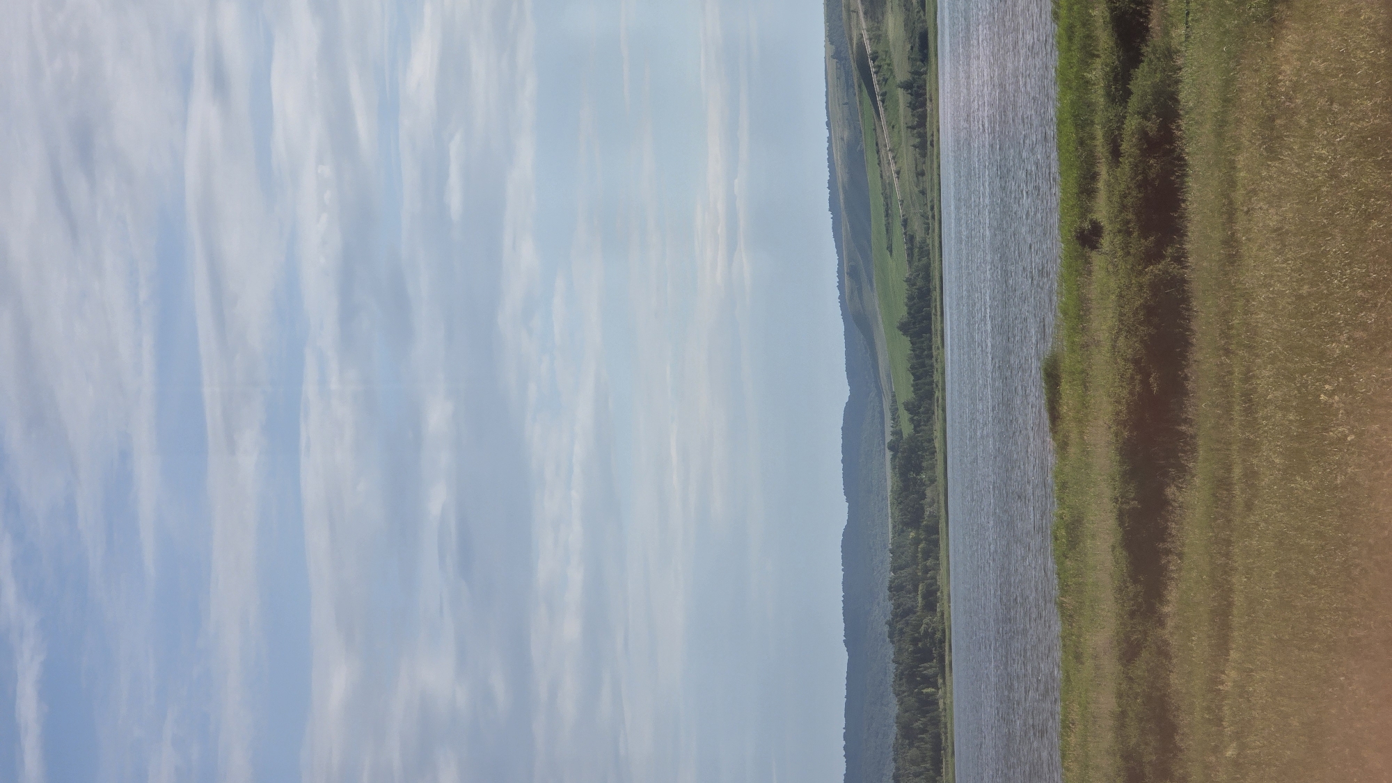 The image depicts a serene landscape scene, likely in or around the village of Kupoyarovo, Russia. The upper portion of the image is dominated by a vast sky with scattered, light-colored clouds against a pale blue background. Below the sky, a gentle expanse of rolling hills is visible, covered in a patchwork of green fields and darker, wooded areas. A body of water, possibly a river or lake, stretches across the middle ground, reflecting the soft light of the sky. In the foreground, a field of tall, dry grass, predominantly golden-brown with some hints of green, slopes gently towards the viewer. The overall impression is one of a tranquil, rural environment on a clear day. There are no people or specific structures visible in the scene.