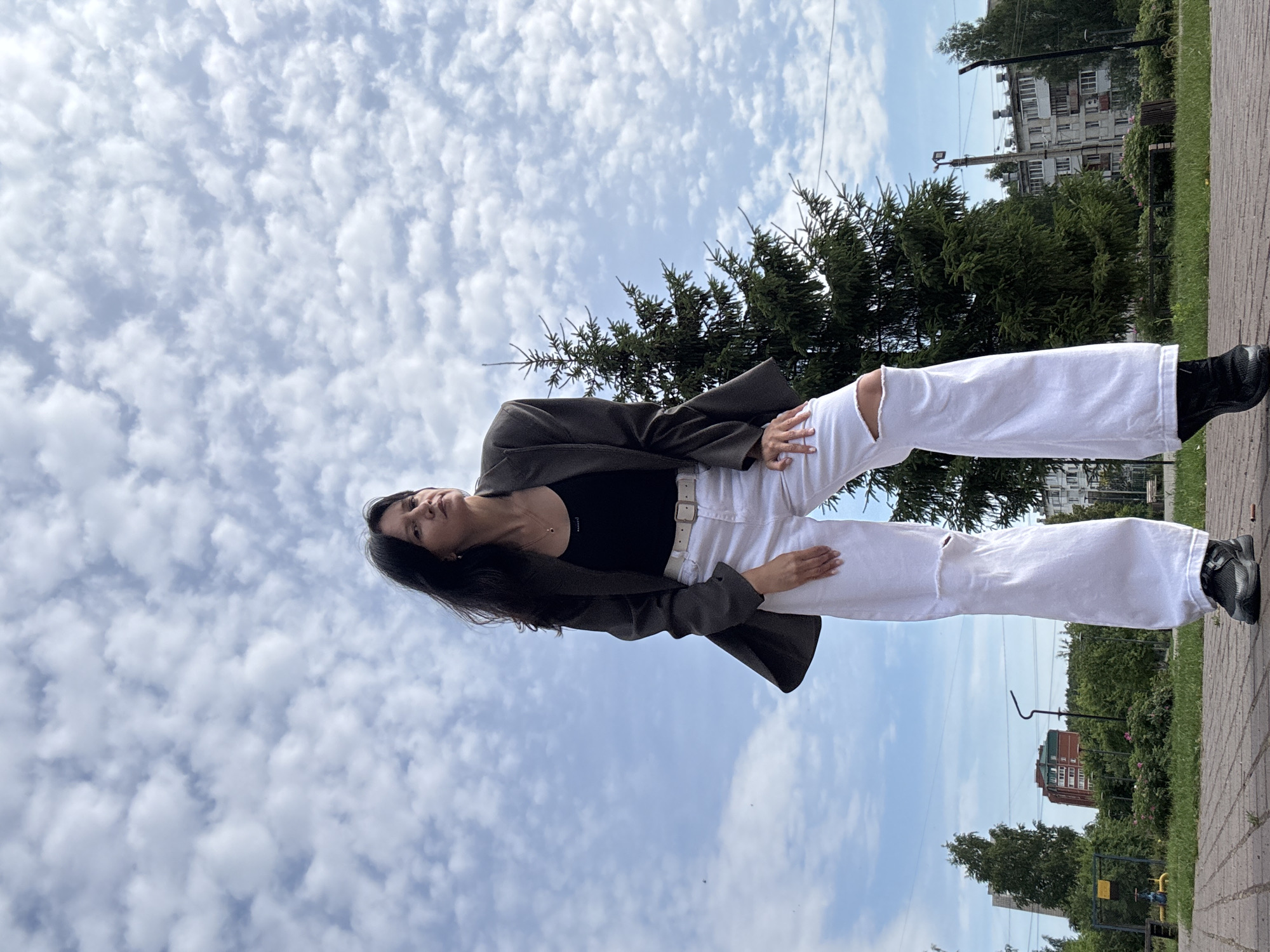 A woman with dark hair, wearing a dark tank top, a dark oversized blazer, and ripped white wide-leg jeans, stands on a paved walkway. She has one hand on her hip and the other resting on her thigh. Behind her, a large evergreen tree stands tall against a cloudy blue sky. In the background, a residential building with multiple stories and a few smaller trees can be seen. The scene appears to be a public outdoor space, possibly a park or a courtyard, within the city of Chaikovsky, Russia.