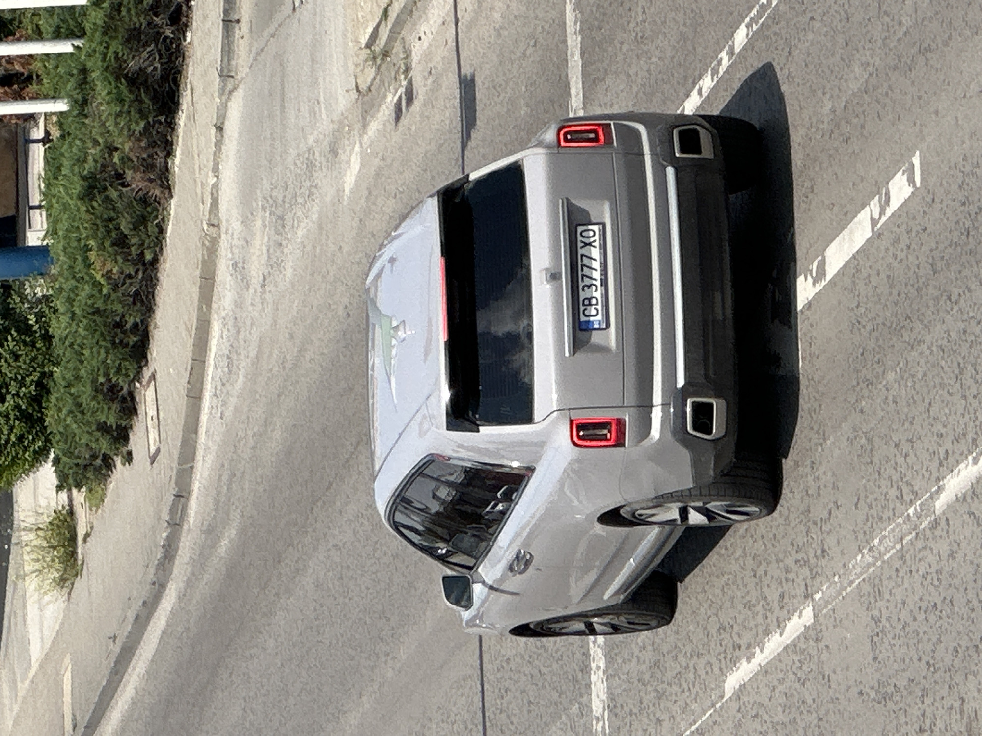 The image, taken from an elevated perspective, captures a light grey modern car, likely an SUV or crossover, parked on an asphalt road. The car is oriented diagonally, with its rear facing the upper right of the frame. Distinctive features of the car include its rectangular LED taillights and a square exhaust outlet visible on the right side of the rear bumper.
The license plate on the rear of the vehicle is clearly visible, displaying the text "CB 3777 XO". The "CB" designation on a Bulgarian license plate indicates Sofia, though the overall location context provided for the image is Veliko Tarnovo, Bulgaria.
The scene is brightly lit by direct sunlight, casting long, dark shadows from the car onto the road, suggesting it is midday or early afternoon on a clear, sunny day. The asphalt road features faded white dashed lines, possibly marking parking spaces or lanes. To the left of the road, a concrete sidewalk is bordered by a lush line of green bushes or hedges, beyond which parts of a building are faintly discernible. No people or other vehicles are visible in the frame, indicating a quiet street or parking area. The car appears to be stationary, likely parked.