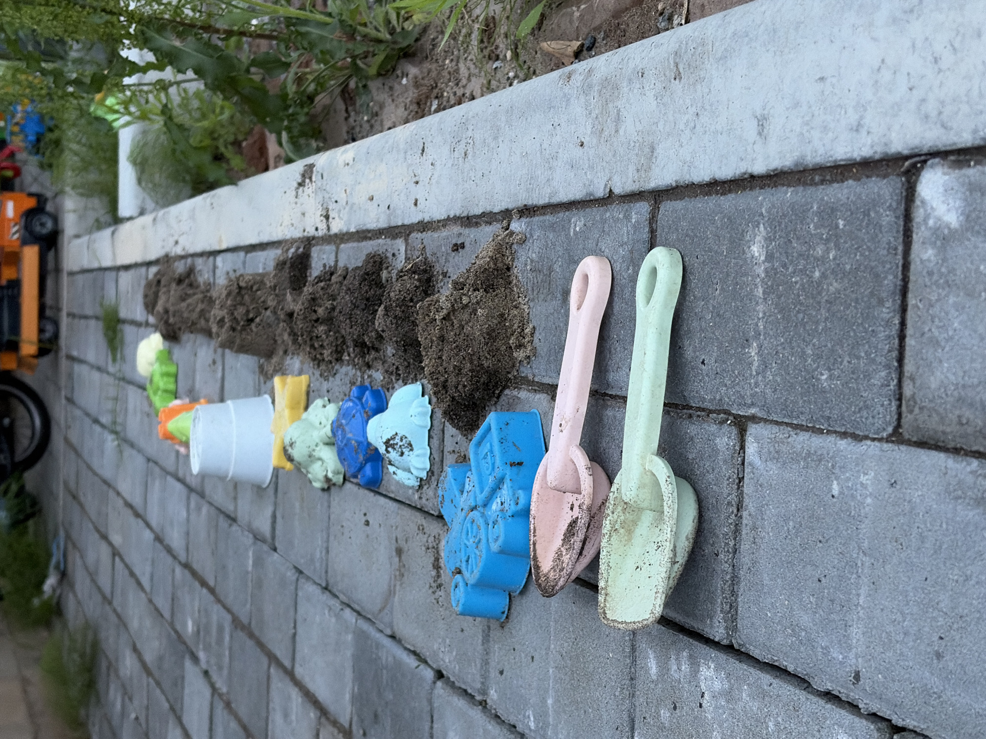 The scene is an outdoor ground-level view in the city of Chaikovsky, Russia, depicting a paved area alongside a dirt and vegetated strip. In the foreground on the left, a light green plastic sand shovel and a light pink plastic sand shovel, both soiled with dark earth, rest on grey rectangular concrete pavers. Behind them, a line of plastic sand molds, also soiled, includes a bright blue vehicle-shaped mold, a light blue turtle mold, a light green amphibian-shaped mold, a yellow rectangular mold, an inverted white plastic bucket, an orange mold, and two additional green and light yellow/green molds further back. To the right of these toys, a row of approximately 10-12 small, conical piles of dark brown soil or sand extends along the edge of the paved path. A grey concrete curb separates the paved area from an adjacent strip of exposed dark soil populated by various green plants and weeds. In the blurred background, green vegetation, a partial dark object (possibly a tire), and an orange toy truck are visible, suggesting a wider outdoor play or storage area. No individuals are present. The arrangement of toys and soil piles indicates recent children's play activities.