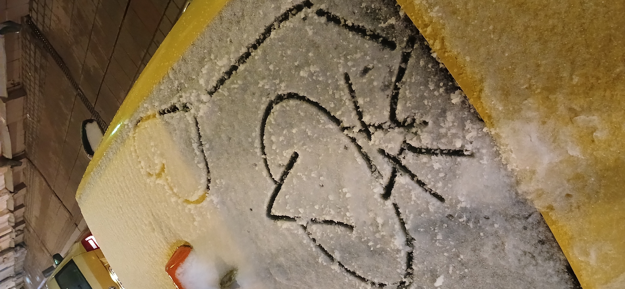 The image depicts a section of a yellow car covered in snow, parked on a street in Moscow, Russia. The snow has been disturbed, revealing a dark surface beneath, on which graffiti-style lettering has been drawn. The environment appears to be urban, with a brick wall visible in the background.