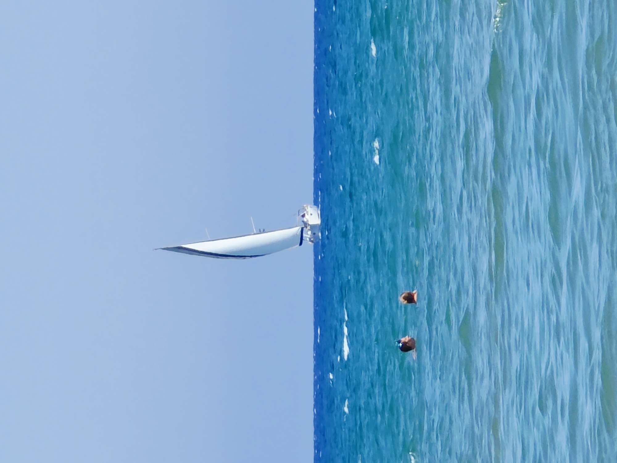 In the sea off the coast of Larnaca, Cyprus, two individuals are submerged in the turquoise waters, their heads and upper shoulders visible above the surface. One person, on the left, wears goggles. In the distance, a white sailboat with a dark sail navigates the blue sea under a clear sky. The horizon is sharply defined between the water and the sky.