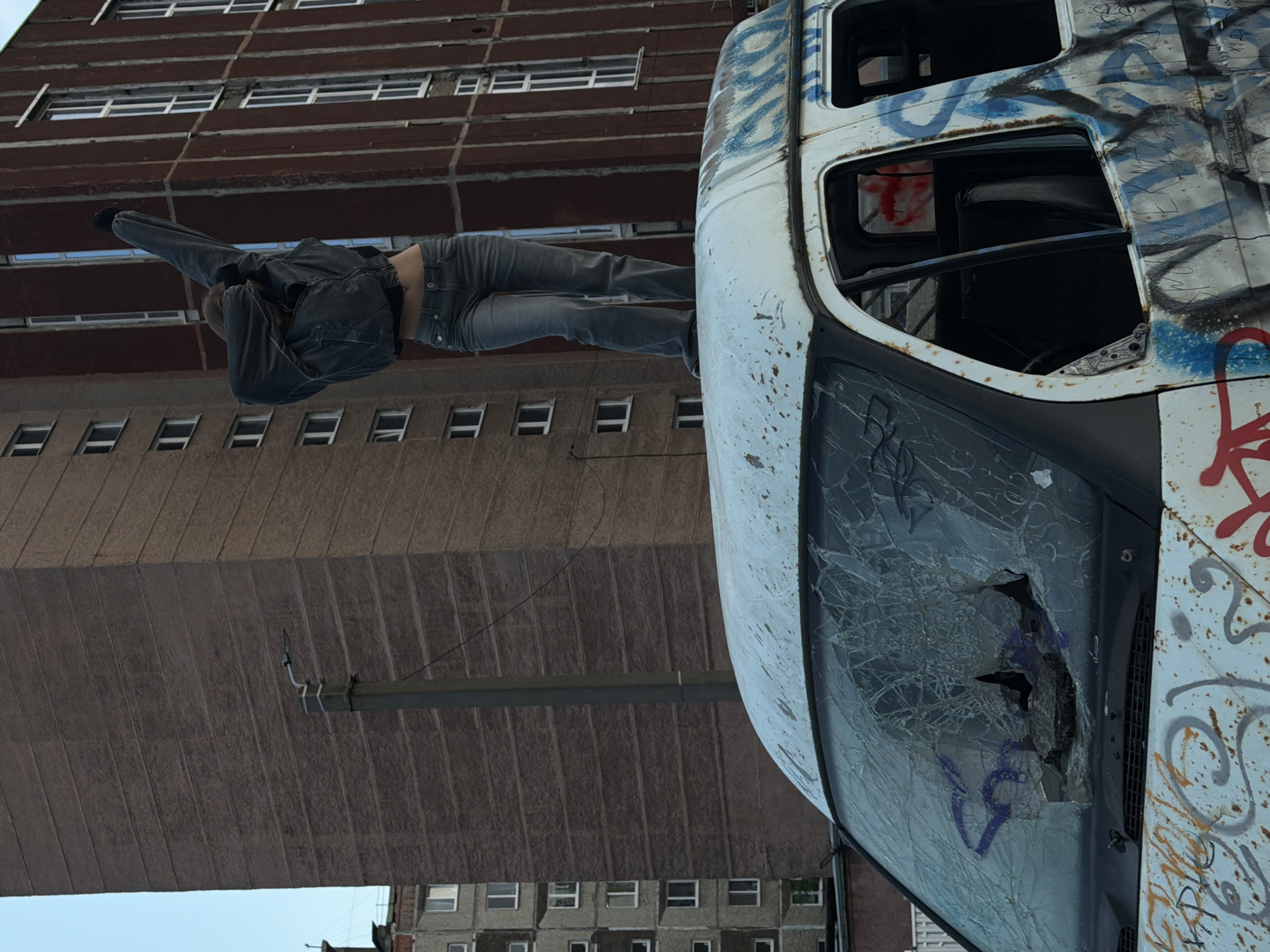 A person stands on the roof of a white, graffiti-covered van with a shattered windshield. The van is positioned in an urban environment, with two large, imposing apartment buildings in the background. One building, on the left, is a light-colored, multi-story structure with many windows. The other, on the right, is a darker, reddish-brown building with vertical window arrangements. A tall, dark lamppost stands between the buildings. The sky above is clear and blue. The person on the van is wearing a denim jacket and jeans, with their arms raised in a dynamic pose. The overall scene suggests a gritty, urban setting, possibly in a post-industrial area. The graffiti on the van and the damaged windshield indicate a state of disrepair or perhaps an artistic statement.