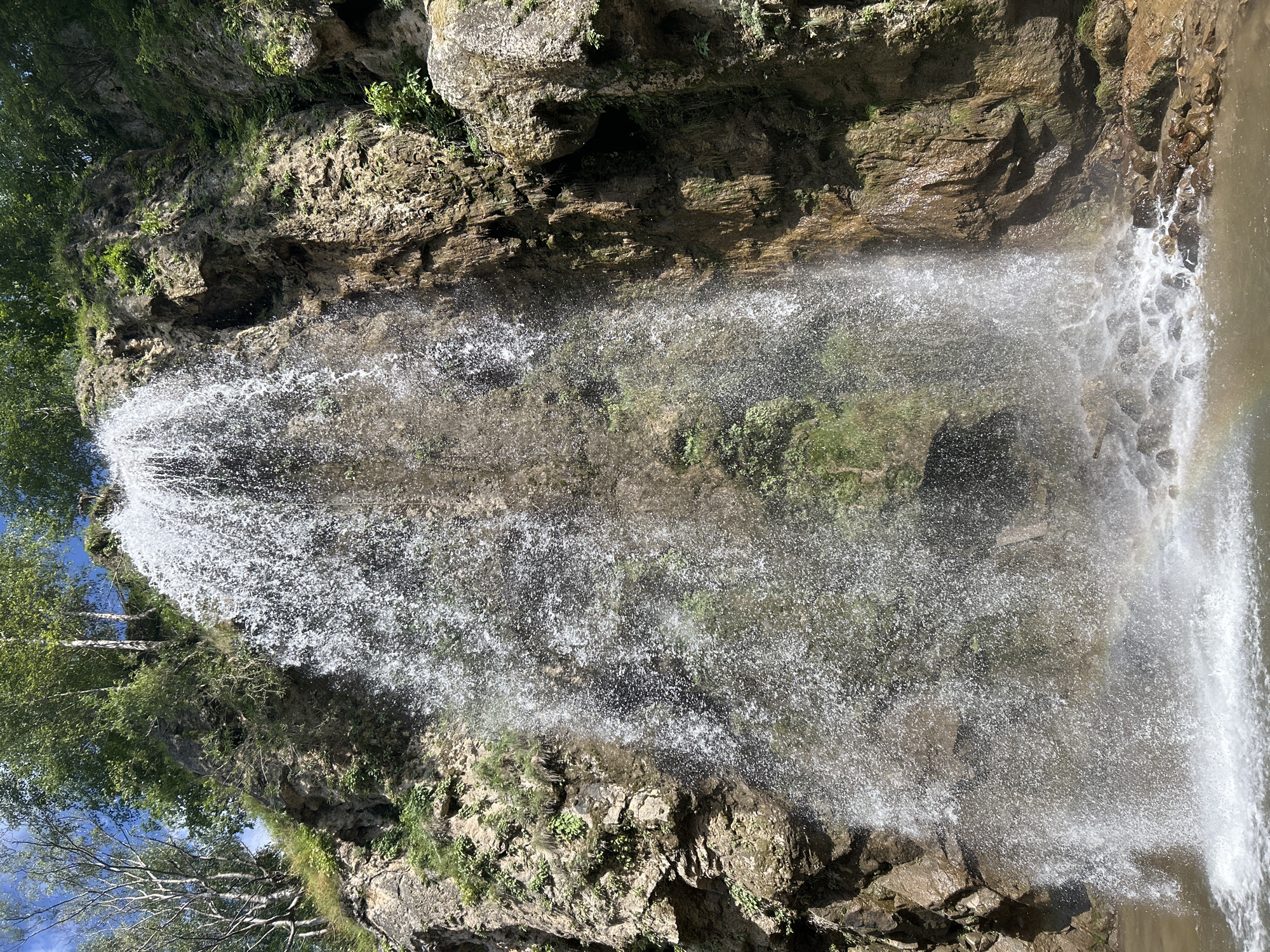 A waterfall cascades down a rocky cliff face covered in green vegetation and trees. The water crashes into a pool at the bottom, creating a misty spray. Sunlight shines through the water, forming a faint rainbow. The rocks are rough and textured, with crevices and ledges. The surrounding area is lush and natural, with a clear blue sky visible above. This scene is located in the city of Krasny Kurgan, Russia.