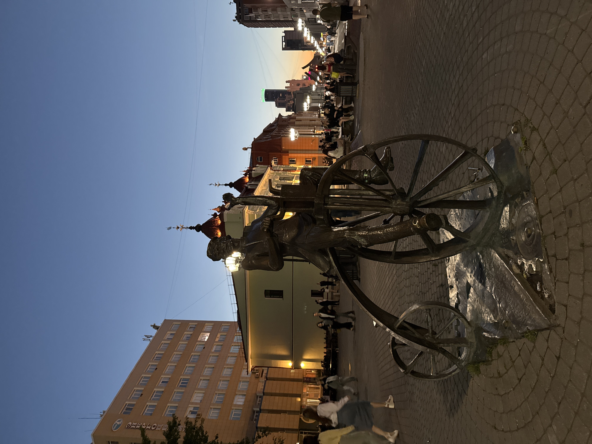 A bronze sculpture of a man riding a penny-farthing bicycle is positioned in the foreground of a city street scene at dusk in Yekaterinburg, Russia. The sculpture is detailed, depicting the man in period clothing, leaning forward as if in motion. The penny-farthing's wheels are large and ornate, with thin spokes. The sculpture is mounted on a textured base that blends into the brick-patterned paving stones of a pedestrian area.
In the midground, a street lined with buildings stretches into the distance under a darkening sky. The buildings vary in architectural style, with some featuring ornate roofs and classical facades, while others are modern high-rises. Streetlights are illuminated, casting a warm glow on the scene. Several people are visible walking and sitting on benches along the street, enjoying the evening.
To the left of the sculpture, a large, modern building with many windows stands, its facade lit by the remaining daylight. On the right, the street curves slightly, with more people and shops visible. The sky is a gradient of deep blue at the top, transitioning to lighter shades of orange and yellow near the horizon, indicating sunset or twilight. The overall impression is of a lively urban environment transitioning from day to night.