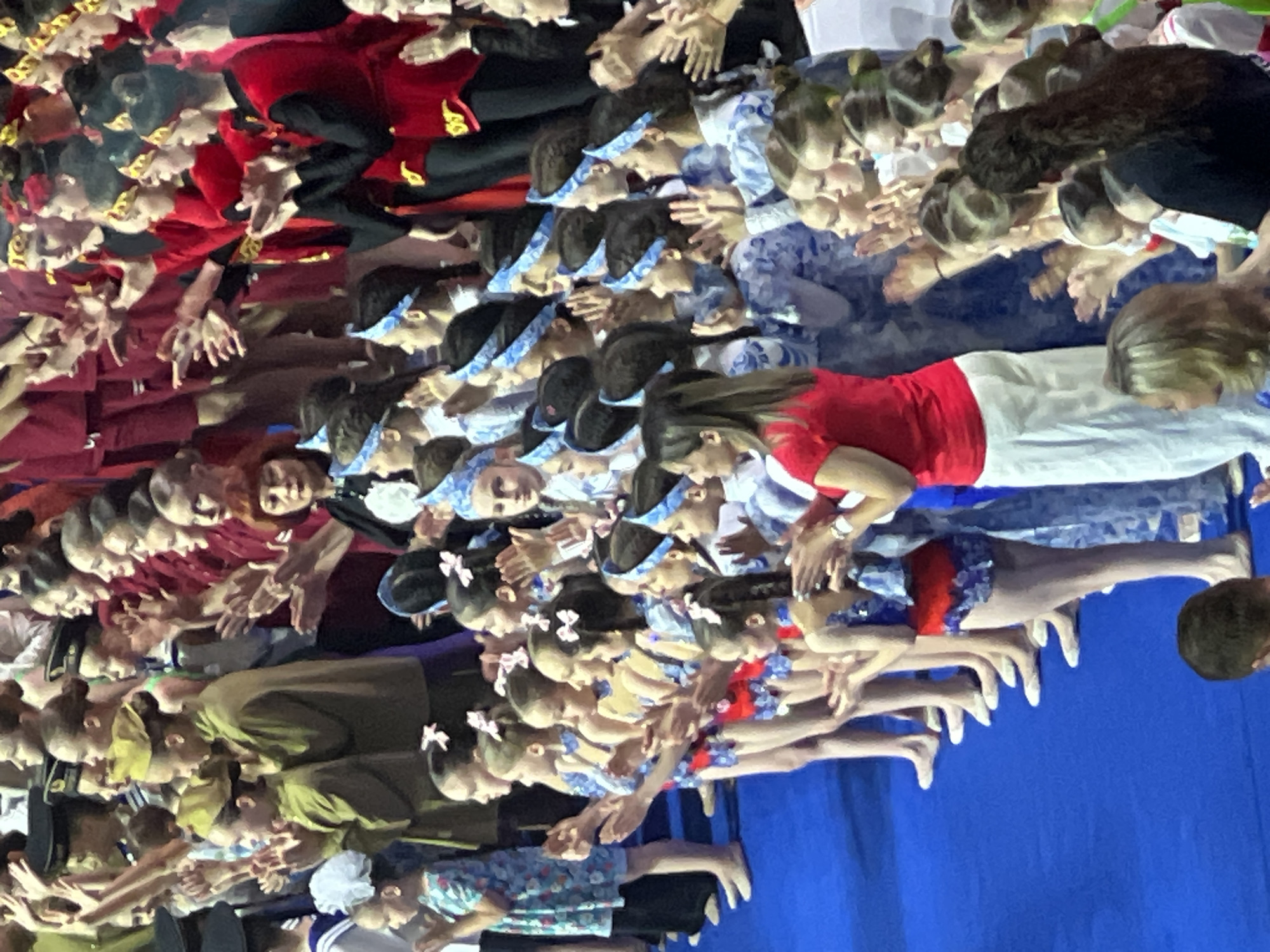 The image depicts a group of young girls in costumes, appearing to be performers, possibly in a gymnastics or dance event. They are standing in rows on a blue surface, some with hands clasped or raised, suggesting they are applauding or performing. Many of the girls are wearing matching outfits, and some have decorative headbands or hair accessories. The lighting suggests an indoor venue. The presence of what appears to be a backdrop or stage setting is implied by the arrangement of people and the overall context of a performance.