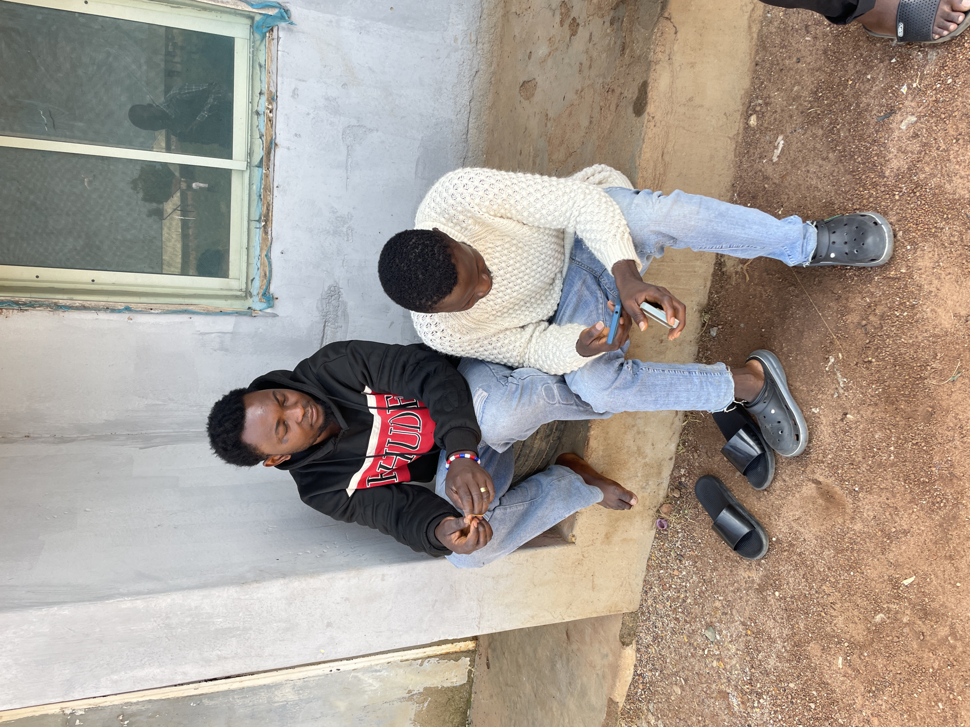 Two young men are sitting on a concrete step outside a building with a rough concrete wall and a dirty, grime-covered window. The man on the left wears a black hoodie with red and white lettering and light blue jeans. He is looking down at his hands, which are clasped together. He is barefoot. The man on the right wears a white, cable-knit sweater and light blue jeans. He is looking at his phone, holding it in his right hand. There are three pairs of casual footwear on the ground in front of the men: a pair of black slides, another pair of black slides, and a pair of black Crocs. The ground in front of the step is a reddish-brown dirt with small stones. The overall environment appears to be somewhat unkempt, possibly an unfinished or neglected outdoor area in Durumi, Nigeria.
