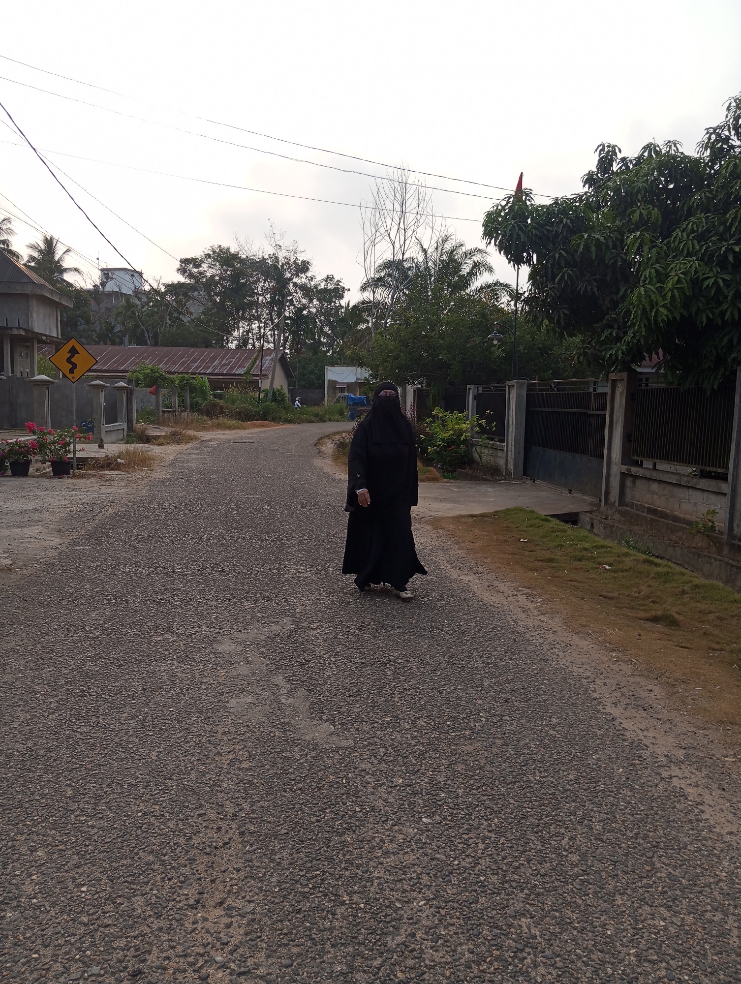 A person wearing a black niqab and abaya walks on an asphalt road in Pematang Reba, Indonesia. The person is positioned in the middle right of the frame, facing left. The road has a rough texture with visible gravel. To the left of the road, there are two potted plants with pink flowers, a yellow zigzag warning sign, a concrete fence, and a building with a corrugated metal roof. Beyond the fence, there are more buildings and trees. To the right of the road, a dark metal fence runs along the property line, with a grassy verge and a concrete drainage ditch. In the background, there are more trees, power lines, and a cloudy sky. A red flag is visible atop a tree in the far right background. The lighting suggests it is either early morning or late afternoon.