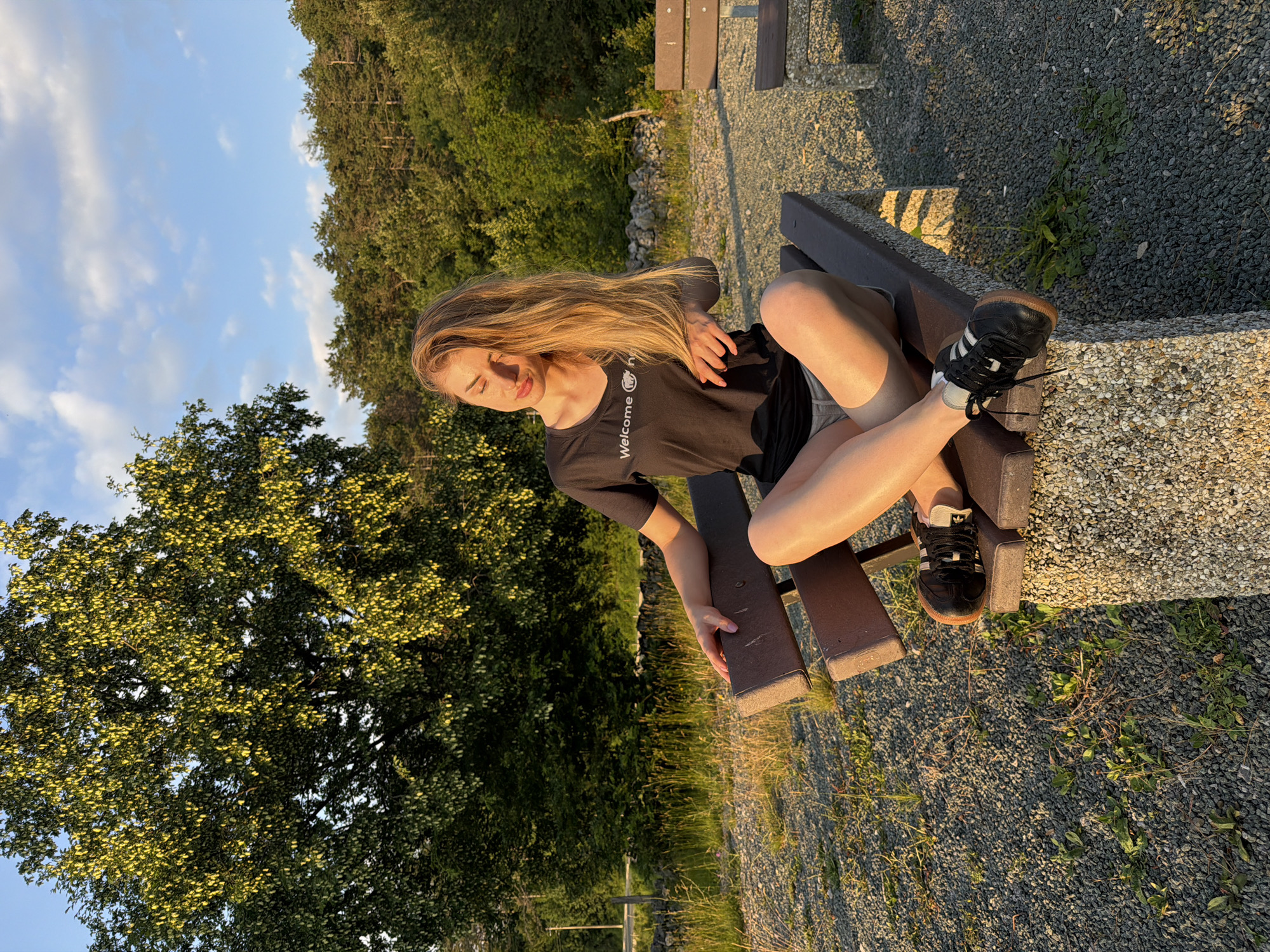 An outdoor, eye-level, medium shot shows a young woman with long blonde hair sitting on a bench in a rural setting, with a backdrop of trees. The woman, facing left, has her eyes closed and her head tilted back slightly, basking in sunlight. She is wearing a black t-shirt with "Welcome" printed on it, grey shorts, and black sneakers with white laces. Her right hand rests on the bench beside her, and her legs are crossed. The bench is made of wood with concrete supports. The ground is covered in gravel and scattered green weeds. In the background, a large tree with green leaves is to the left, and a dense forest is visible further back. The sky above is blue with scattered clouds, and the sunlight casts long shadows. The scene appears to be in a park or natural area with a few other benches visible in the distance.