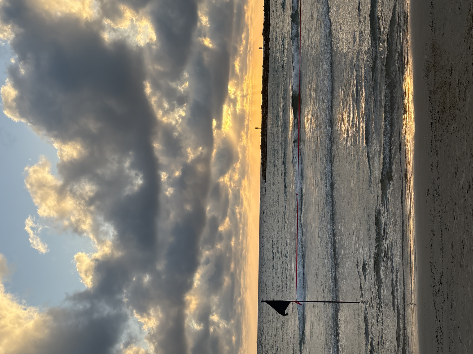 This is a vertical, eye-level shot of a beach scene at sunset. The image is divided into two distinct halves by the horizon line. The left side of the frame is dominated by a dramatic sky filled with a mix of dark, heavy clouds and lighter, golden-tinged clouds, suggesting the sun is low in the sky and breaking through. The sky transitions to a soft, warm yellow near the horizon.
The right side of the frame depicts the ocean and the beach. The water is a calm expanse with gentle waves reflecting the warm light of the sunset, creating a textured, silvery surface. A thin red rope is strung between two poles, marking a boundary or perhaps a net. In the foreground, a small black flag flies from a pole planted in the sand. The sand itself is visible on the far right of the frame, appearing pale and smooth, with the light of the setting sun casting a warm glow on its surface. In the distance, along the horizon, there appear to be a few small, vertical structures, possibly lights or buoys. The overall mood is serene and peaceful, capturing the quiet beauty of a beach at dusk. The location is identified as Tel-Aviv, Israel.