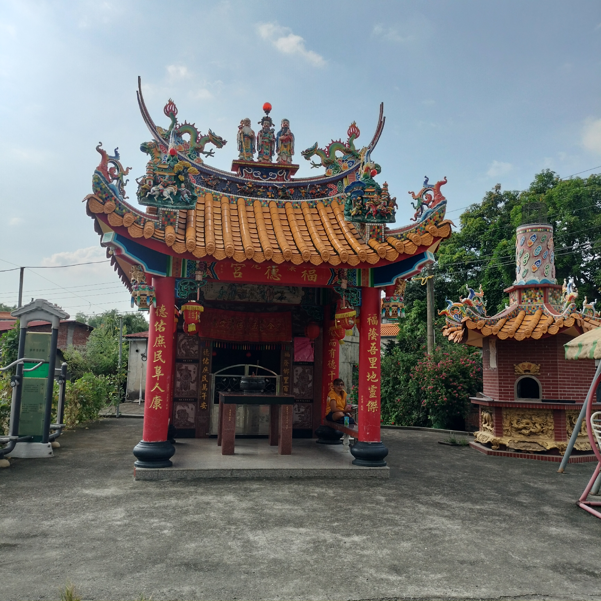 The image shows a brightly colored, ornate temple in Zhushan Township, Taiwan. The temple features intricate roof decorations with dragon motifs and figures, and its pillars are inscribed with red and gold Chinese characters. In front of the main temple structure is a low altar with an incense burner. To the right of the main temple, there is a smaller brick structure, possibly an incense burning furnace, also adorned with yellow decorative elements and a tiled roof. A person wearing a yellow shirt and dark shorts is seated on a bench to the right of the main temple, looking towards the right side of the frame. The ground in front of the temple is paved with concrete. Lush green trees and some flowering bushes are visible in the background, along with a clear sky with scattered clouds. A signpost with green panels is visible to the left of the temple. The overall scene depicts a religious site in a semi-rural or suburban setting.