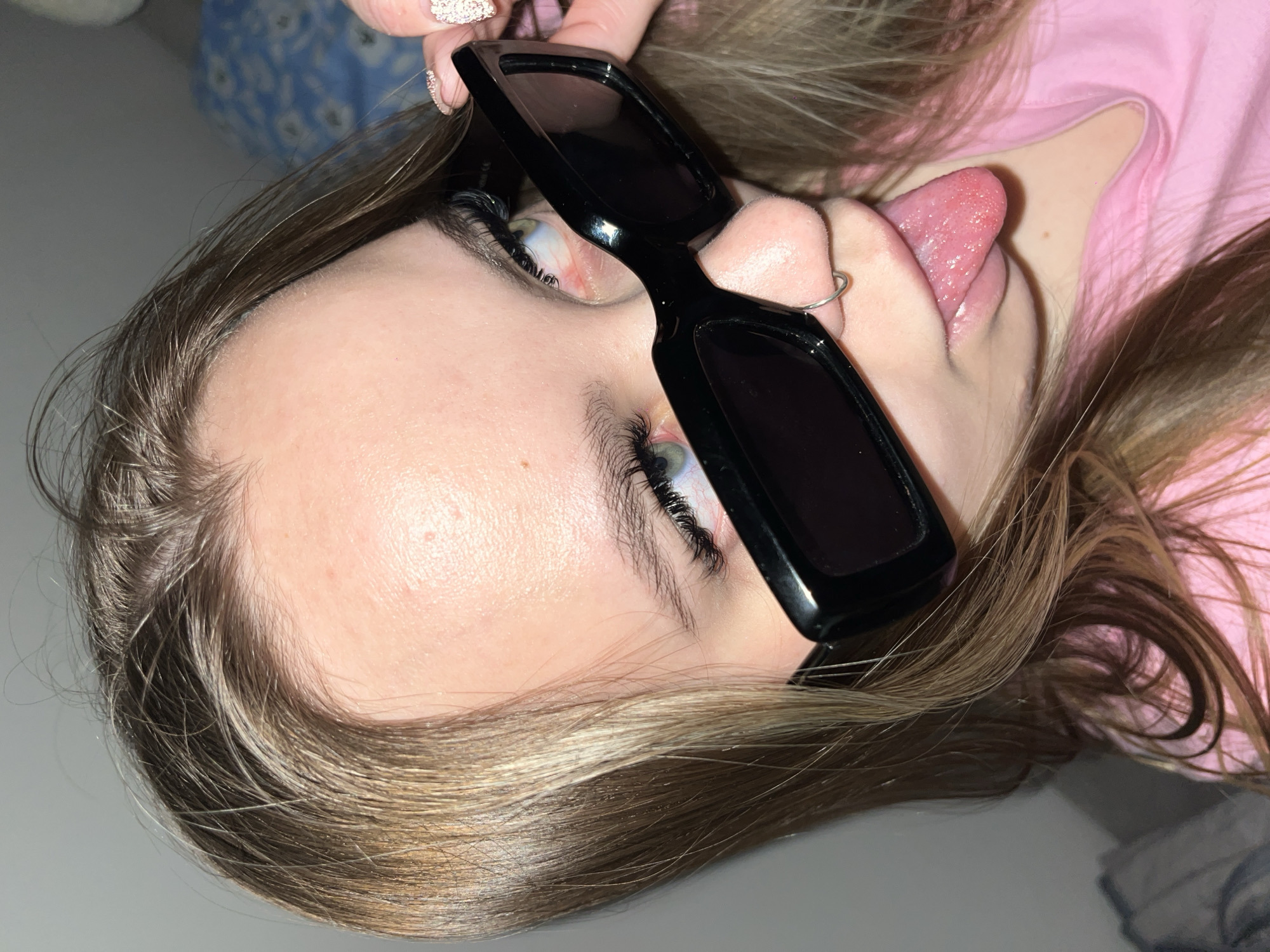 The image is a close-up portrait of a person, likely female, depicted at an angle, with her head tilted, creating a dynamic, rotated composition. She has fair skin, light brown hair with blonde highlights, and striking green-blue eyes. The person is wearing a pink garment, possibly a shirt.
Her right hand, adorned with glittery nail polish, holds a pair of black, rectangular sunglasses over her right eye. Her left eye is open, revealing long, dark eyelashes. A silver nose ring is visible on her nose. In a playful gesture, her tongue is extended out of her mouth, showing its textured surface with visible taste buds.
Her hair, a blend of light brown and blonde, frames her face. The background features blurred elements: a blue and white floral pattern, possibly bedding, in the upper left, and a plain light surface elsewhere. The overall impression is one of casualness and playful expression. There is no visible text. While the location context is Krasnodar, Russia, no specific geographical indicators are present within the visual content of the image itself.