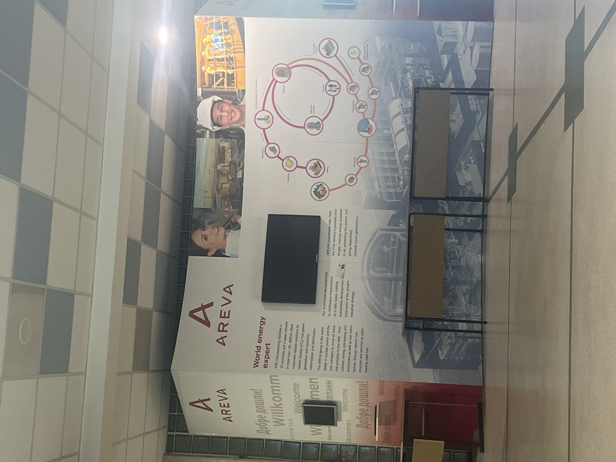 The image shows a display by AREVA, a world energy expert, likely in a commercial or public building in Sofia, Bulgaria. The display includes wall graphics with text in multiple languages, including Bulgarian ("Добре дошли!"), and imagery related to energy production. A black screen and two chairs are positioned in front of the display. The environment suggests a modern interior with tiled walls and flooring.