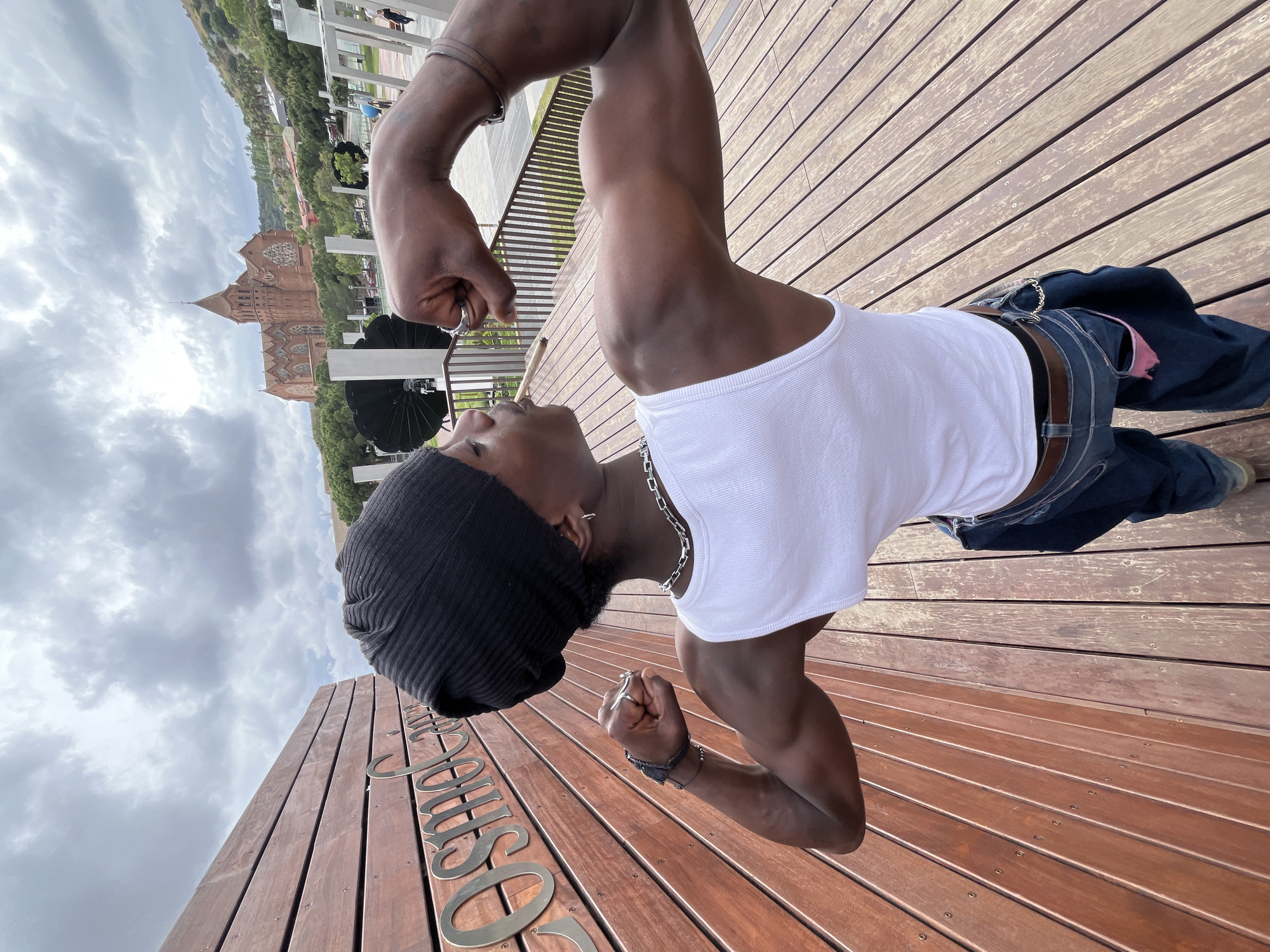 A man is standing on a wooden deck, flexing his arms. He is wearing a white tank top, dark jeans with a brown belt, and a black beanie. He has a silver chain around his neck and bracelets on his wrists. The deck appears to be part of an outdoor area, and behind him is a view that includes a large, ornate building resembling a cathedral. The sky is cloudy. The name "SOULIS" is written on the wooden deck in front of him. This scene is taking place in Barcelona, Spain.