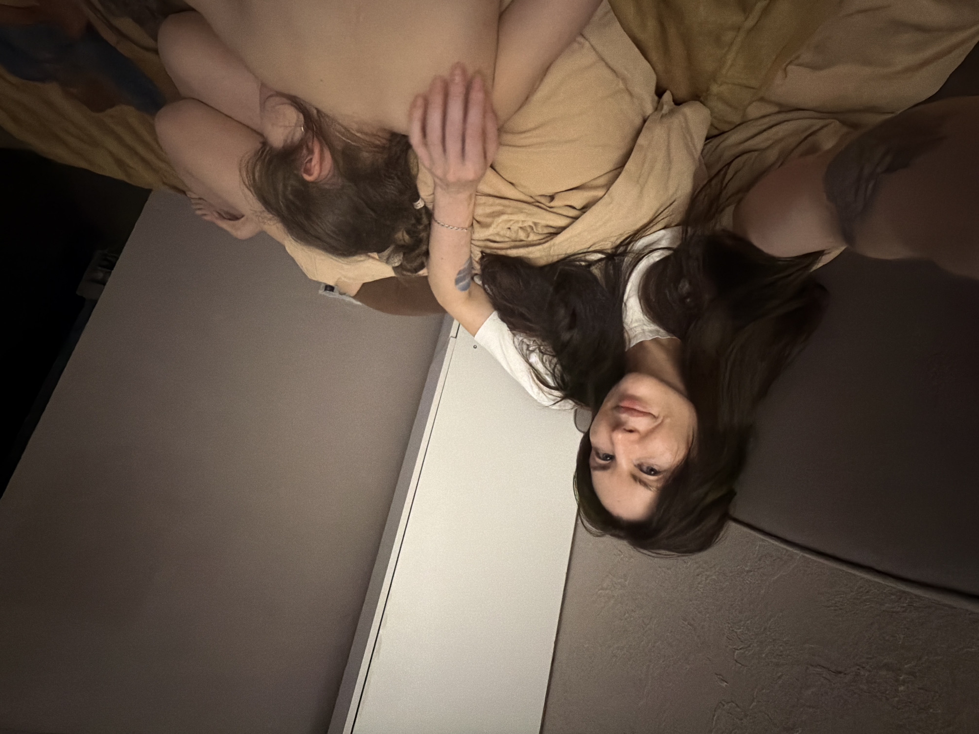 A woman is lying in bed in a room. Another person is lying next to her on their stomach, facing away from the camera. The woman is wearing a white t-shirt and has long brown hair. She is looking at the camera. The room has a dark gray accent wall and a white door frame. The bedding is a light brown color. It is unknown if the location is in Perm, Russia.