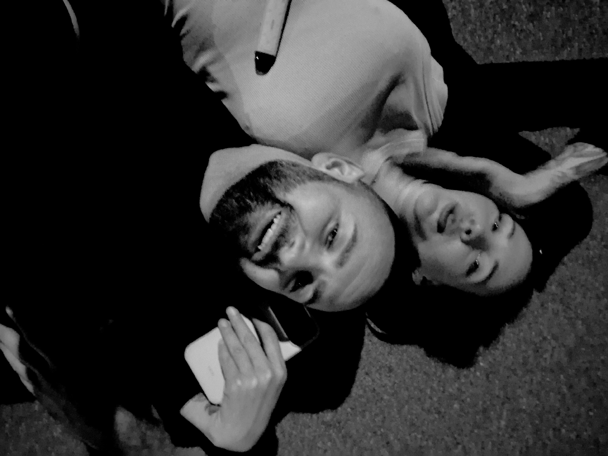 In a low-angle, black and white selfie, a man with a beard smiles widely while holding a smartphone in his right hand. He is lying down, with the woman behind him also lying down. She has her right arm raised and resting on her head, and her mouth is open. The background appears to be a rough, textured surface, possibly pavement or dirt. The overall lighting is dim, creating a contrast between the illuminated faces and the dark clothing and surroundings.