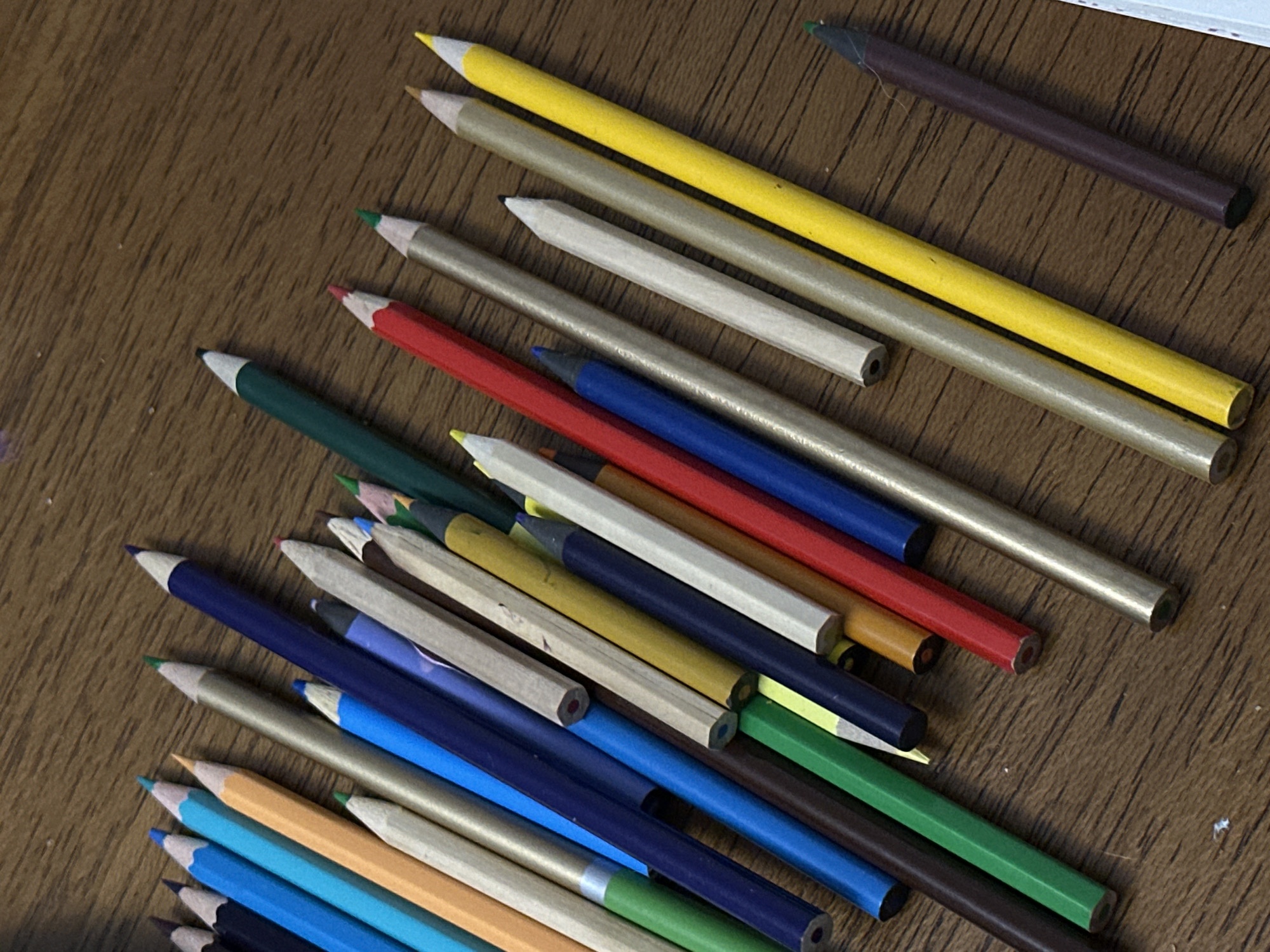 The image depicts a collection of colored pencils scattered on a dark wooden surface. The pencils are in various colors, including yellow, silver, red, blue, green, and brown. The pencils are oriented at various angles, with some overlapping. No people are visible. The setting appears to be indoors. There is no indication of the location of the image being in Tbilisi, Georgia.