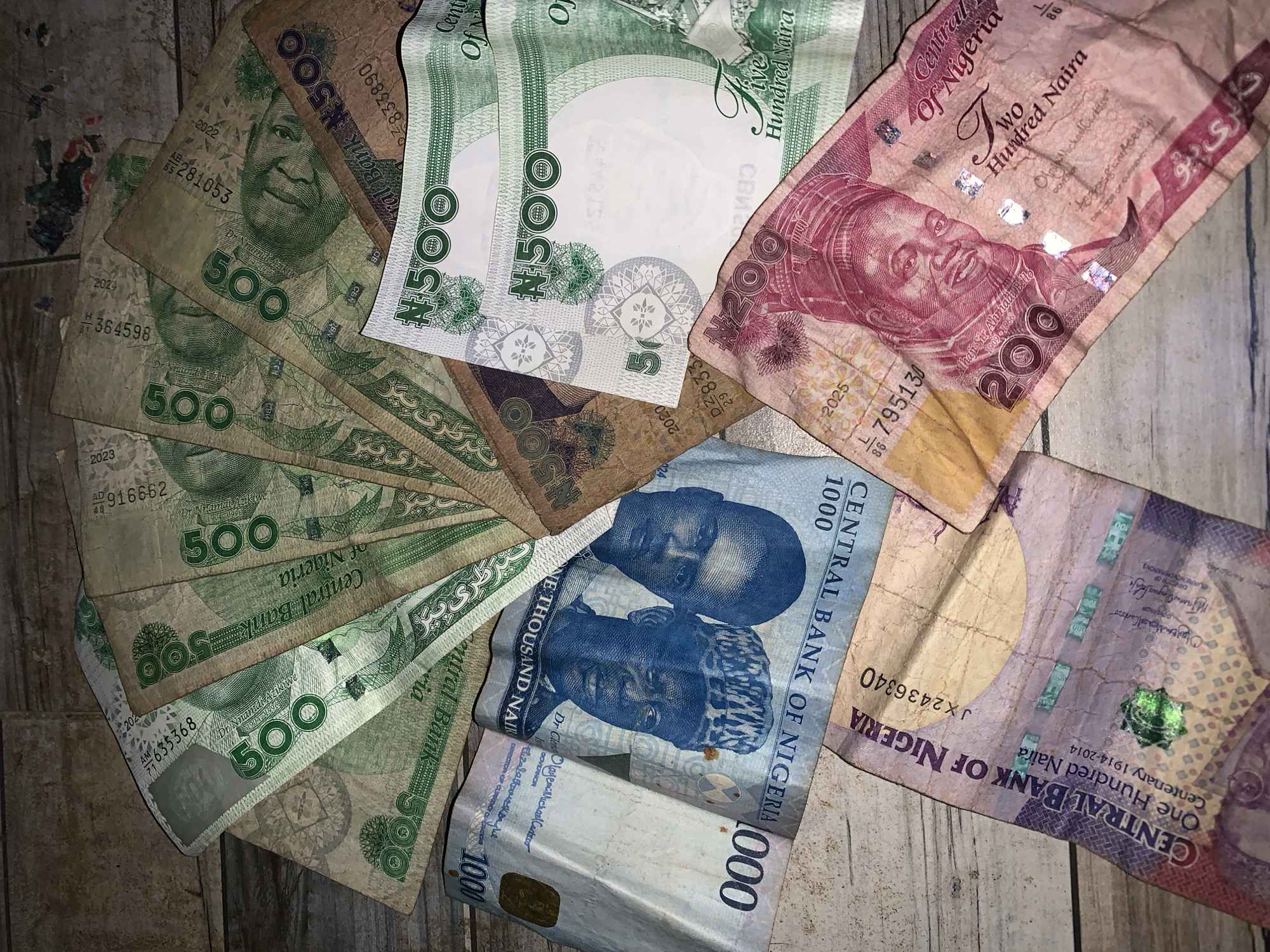 The image shows a collection of Nigerian Naira banknotes arranged on a wooden surface. The denominations visible include ₦500, ₦200, and ₦1000. The banknotes are scattered and partially overlapping. The ₦500 notes feature the portrait of Dr. Nnamdi Azikiwe. The ₦200 note depicts Alhaji Sir Ahmadu Bello. The ₦1000 notes feature two portraits. The environment is a wooden surface. The banknotes are the primary subjects of the image. There is no indication of the location being in Jos, Nigeria.