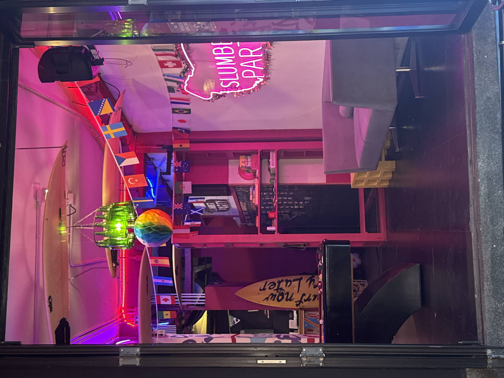 This is an image of the interior of a shop, likely a bar or club, seen from the outside. The shop appears to be decorated with international flags, surfboards, and neon lights. There is a neon sign that reads "SLUMBI PAR" which is likely part of a longer phrase. The lighting is primarily pink and purple, creating a vibrant and festive atmosphere. The scene is well-lit, suggesting it is either daytime or the shop has strong interior lighting. There are no people visible in the image. The overall impression is one of a lively and eclectic space, possibly catering to tourists given the international flags and surfboards. The presence of multiple surfboards suggests a connection to surfing culture, common in beachside locations like Karon, Thailand. There is a small surfboard with the words "surf now party later" written on it in yellow.