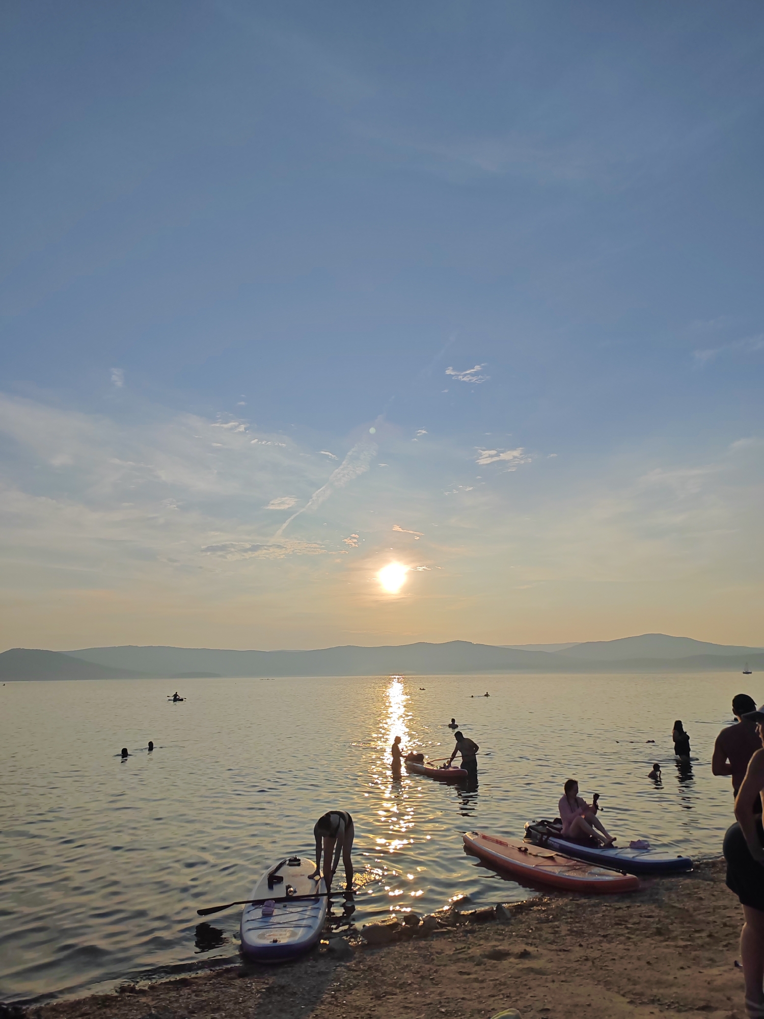 This is an outdoor, full-frame, vertical shot taken at sunset in Turgoyak, Russia. The scene depicts a group of people engaged in water activities on a calm body of water, likely a lake, with rolling hills in the background. The sun is setting, casting a warm, golden light across the water and sky, creating a serene atmosphere. Several individuals are seen paddleboarding, with at least two paddleboards resting on the shore in the foreground. Some people are wading in the shallow water, while others are further out, enjoying the evening. The sky is a gradient of blue and orange, with wispy clouds and contrails visible. The shoreline is covered in pebbles and sand. The overall impression is one of leisure and relaxation during a summer evening.
