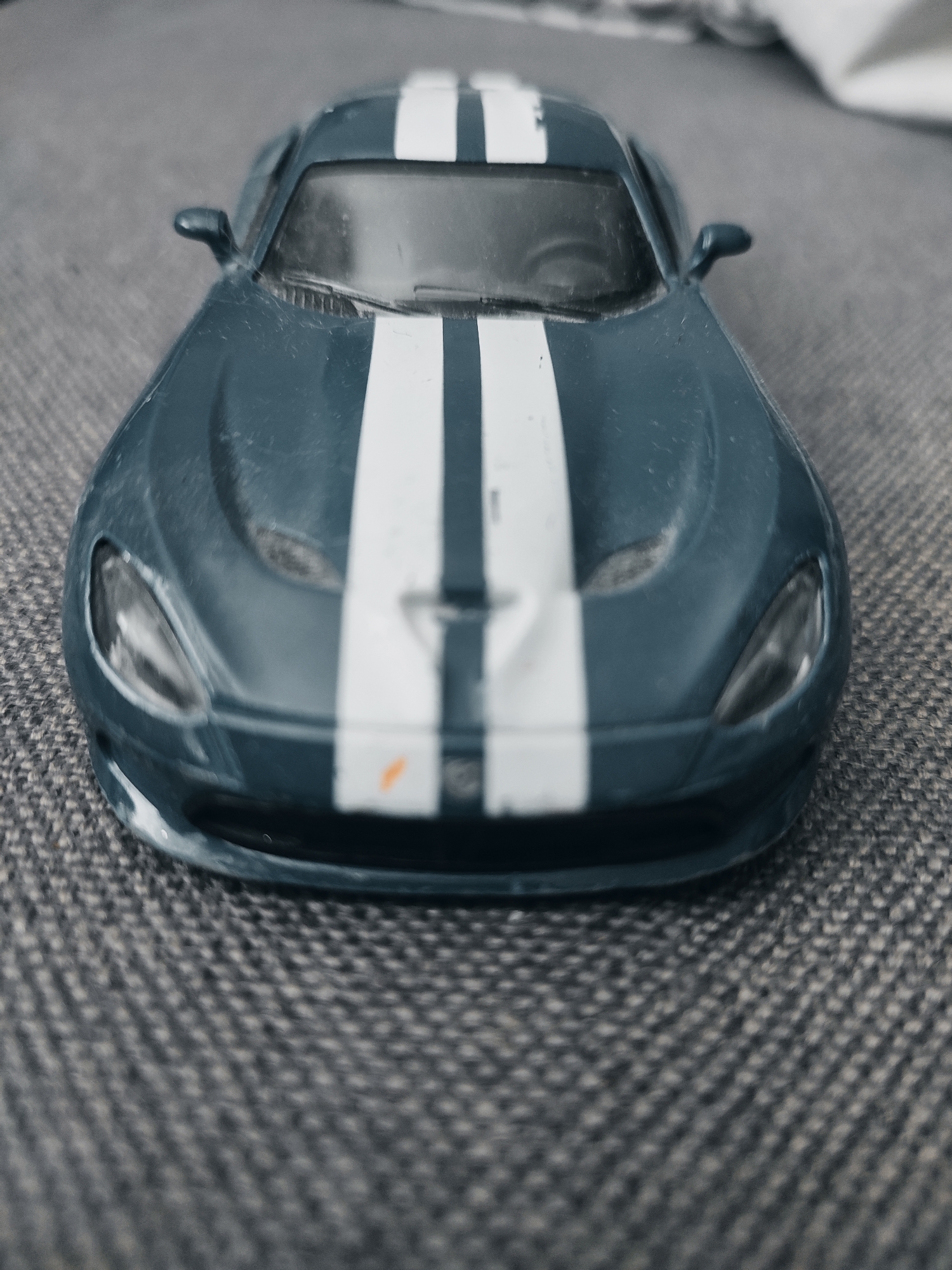 An eye-level, close-up, static shot focuses on the front of a dark blue toy car with white racing stripes. The car is positioned centrally on a textured grey surface that appears to be fabric, possibly upholstery or carpet. The lighting is subdued, casting soft shadows. The car's headlights are visible and reflective. The background is blurred, obscuring details of the surrounding environment. There is no indication of Zlatopil, Ukraine, or any other specific city. The image is devoid of people or any discernible activity.