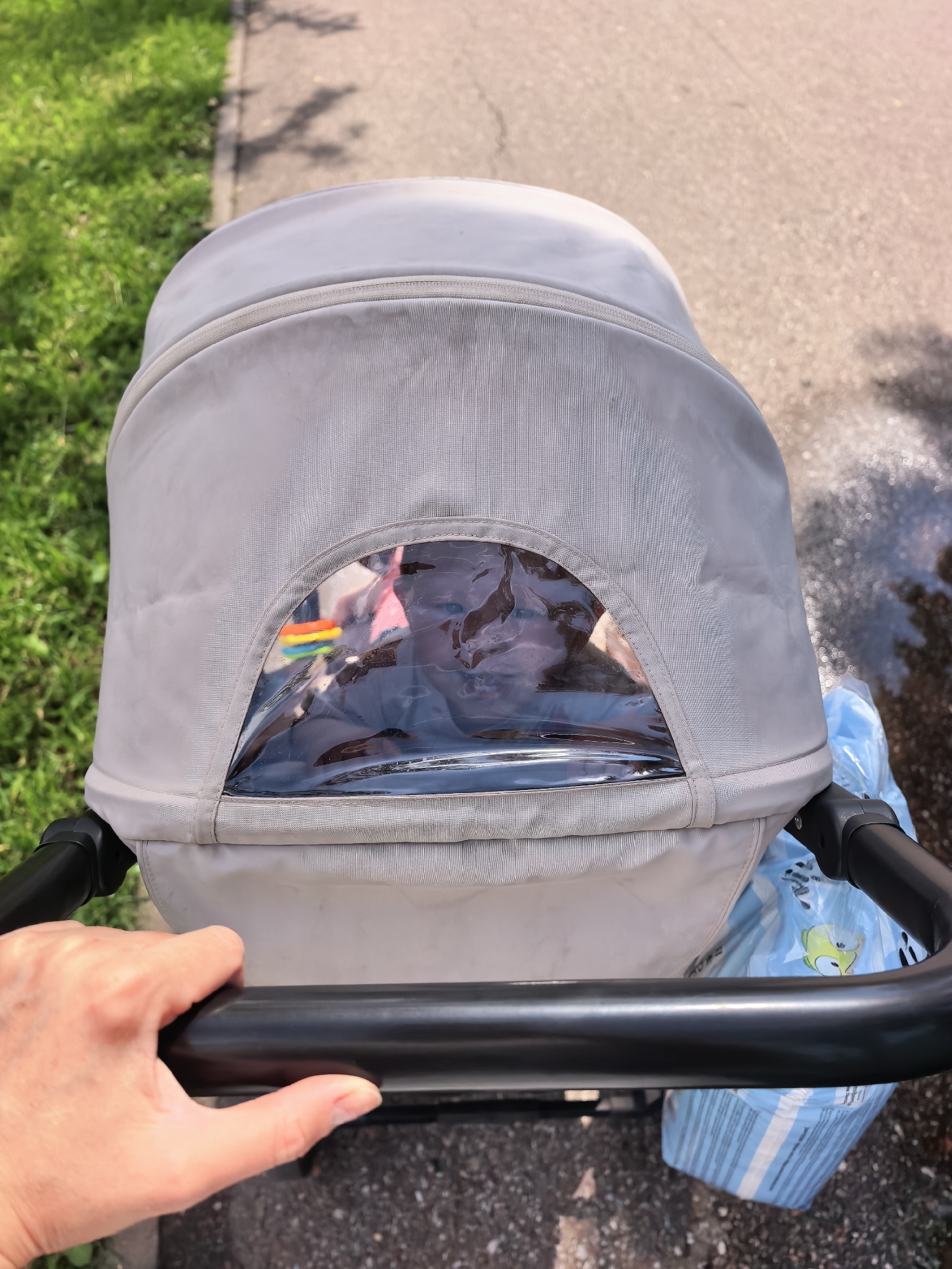 An overhead perspective captures a baby stroller's canopy, which is made of light gray fabric. A clear plastic window is present in the canopy, revealing a baby inside, partially obscured by reflections. A small rainbow-colored object is visible through the window. A person's hand, with lightly tanned skin and visible fingernails, grips the black handle of the stroller. To the right of the stroller, a blue bag with a cartoon character print is partially visible. The stroller is positioned on a paved surface, likely a sidewalk or pathway, with a strip of green grass running along the left edge of the frame. The background shows more pavement with dappled sunlight and shadows. The scene appears to be outdoors, during the day, in a city park or residential area. The location is Sterlitamak, Russia.