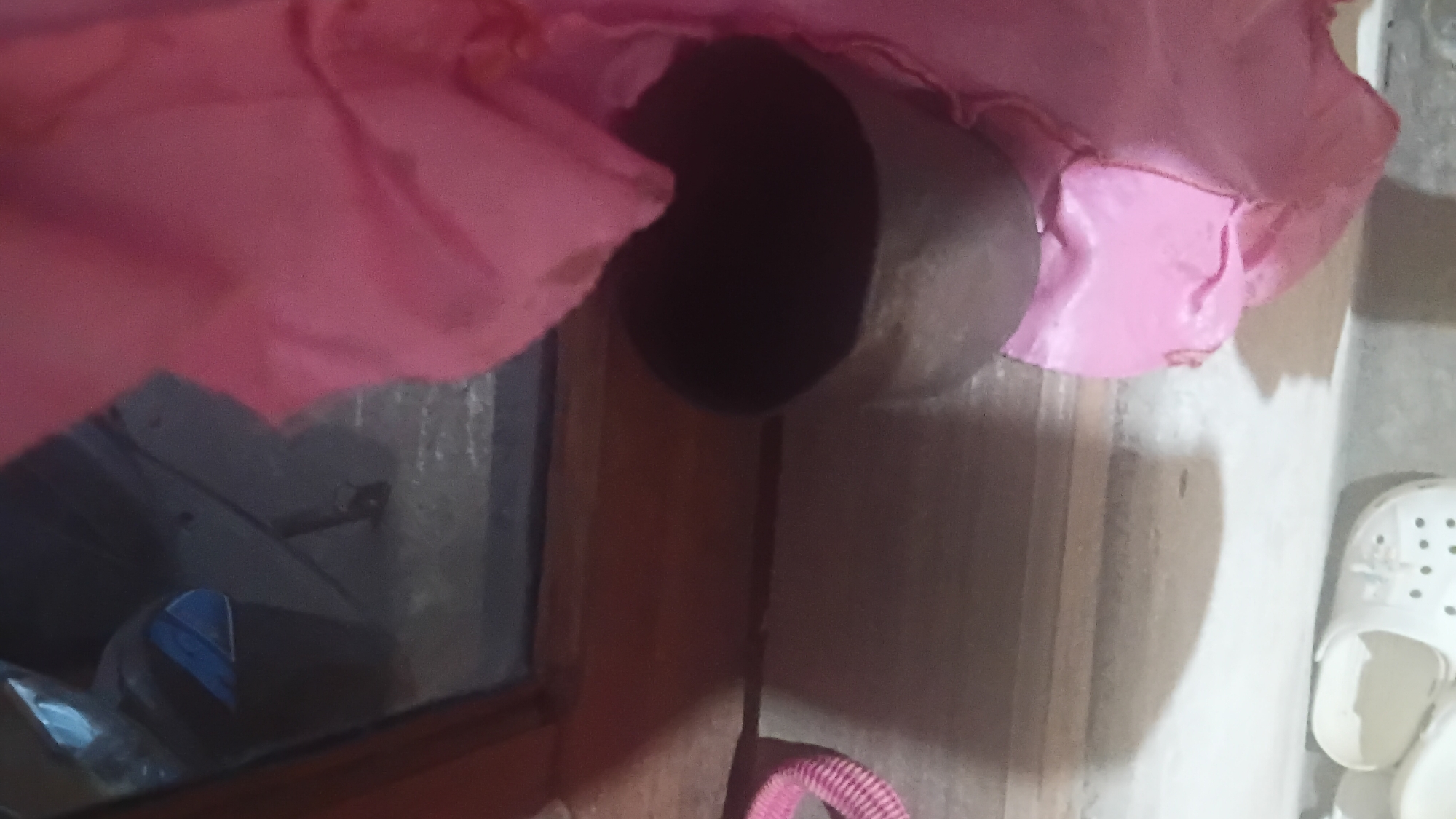A shallow, dark-colored bowl sits on a light-colored floor next to a pink fabric, partially obscuring the bowl. The bowl is positioned in front of a wooden surface, likely a windowsill or shelf, which is in front of a window reflecting a dark, indistinct interior with what appears to be a blue object. To the left of the bowl, a pink ribbed object is visible. In the lower portion of the image, a white shoe with perforated detailing is partially visible on the floor. The scene is indoors, with a focus on everyday objects. The city of Prabumulih, Indonesia, is not depicted or implied in this image.