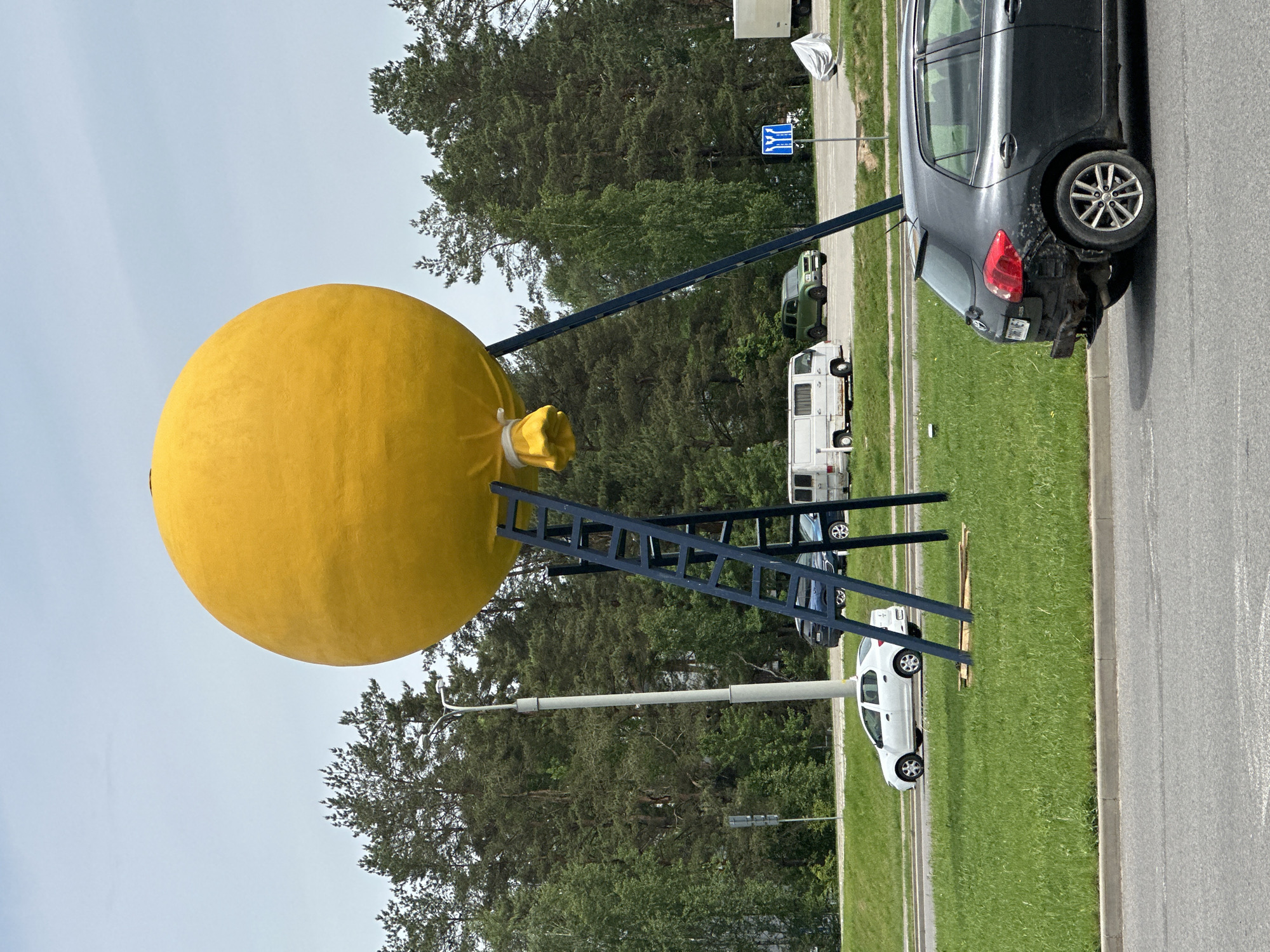 A vertical, outdoor shot features a large yellow sphere sculpture supported by a blue metal frame. The sculpture is located on a grassy area next to a road in Бараўлянскі сельскі Савет, Belarus. Several cars are parked on the side of the road, including a black car with rear damage and a white car. Trees form a backdrop behind the sculpture and cars. A street light is also visible in the background.